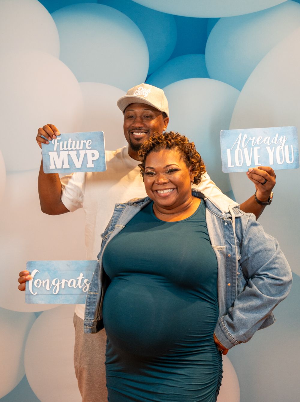 Pregnant couple smiling at baby shower, holding signs reading Future MVP, Already Love You, and Congrats.