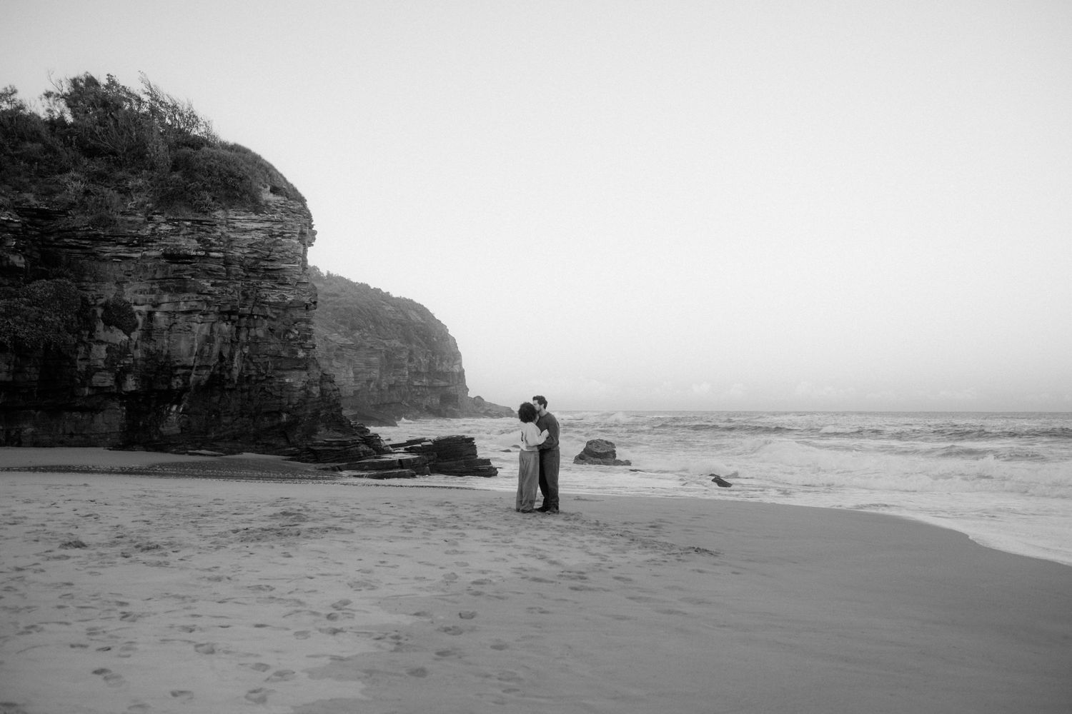 Couple kissing on an empty beach with rocky cliffs behind them.