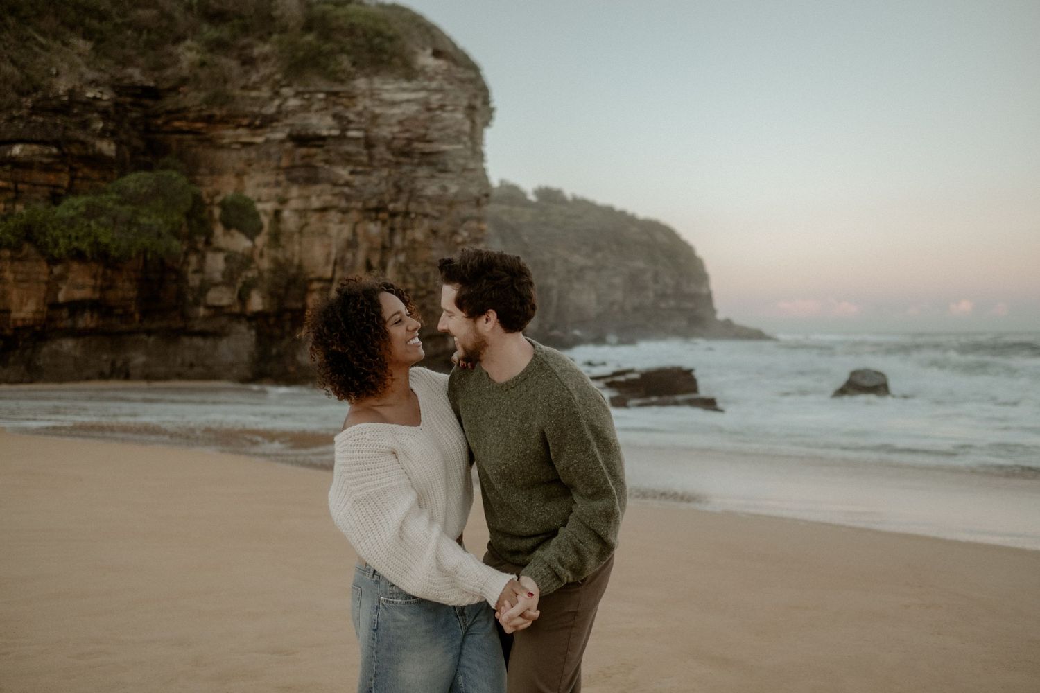 Smiling couple dancing on a sandy beach with cliffs behind them.