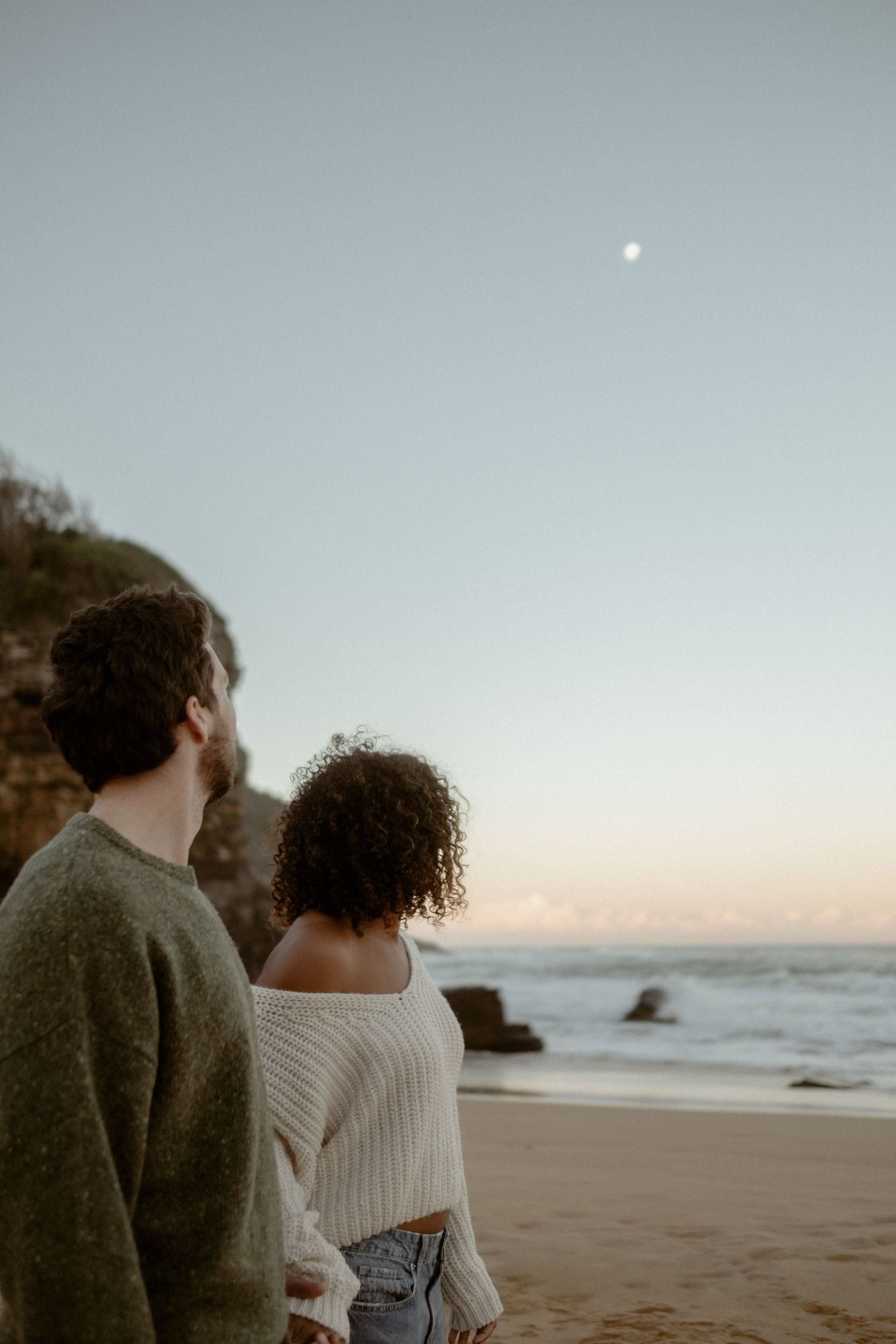 Couple standing on a beach and looking at the moon at sunset.