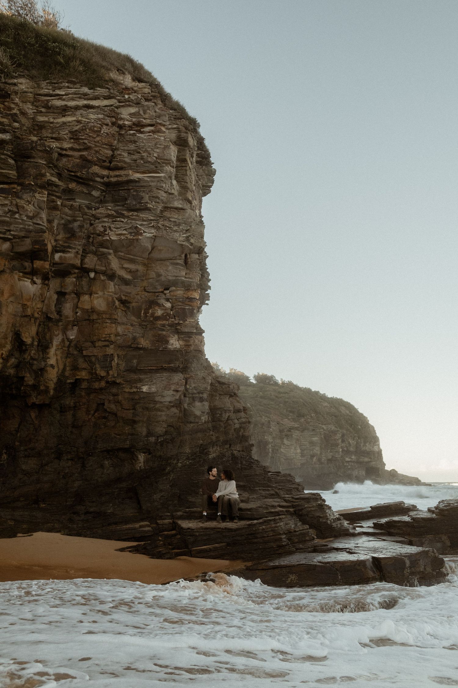 Couple sitting on rocks on a sandy beach with cliffs and. the ocean behind them.