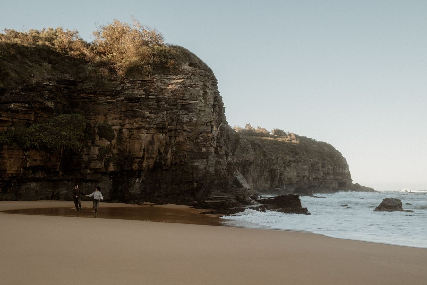 Couple holding hands and running along an empty beach with cliffs behind them.