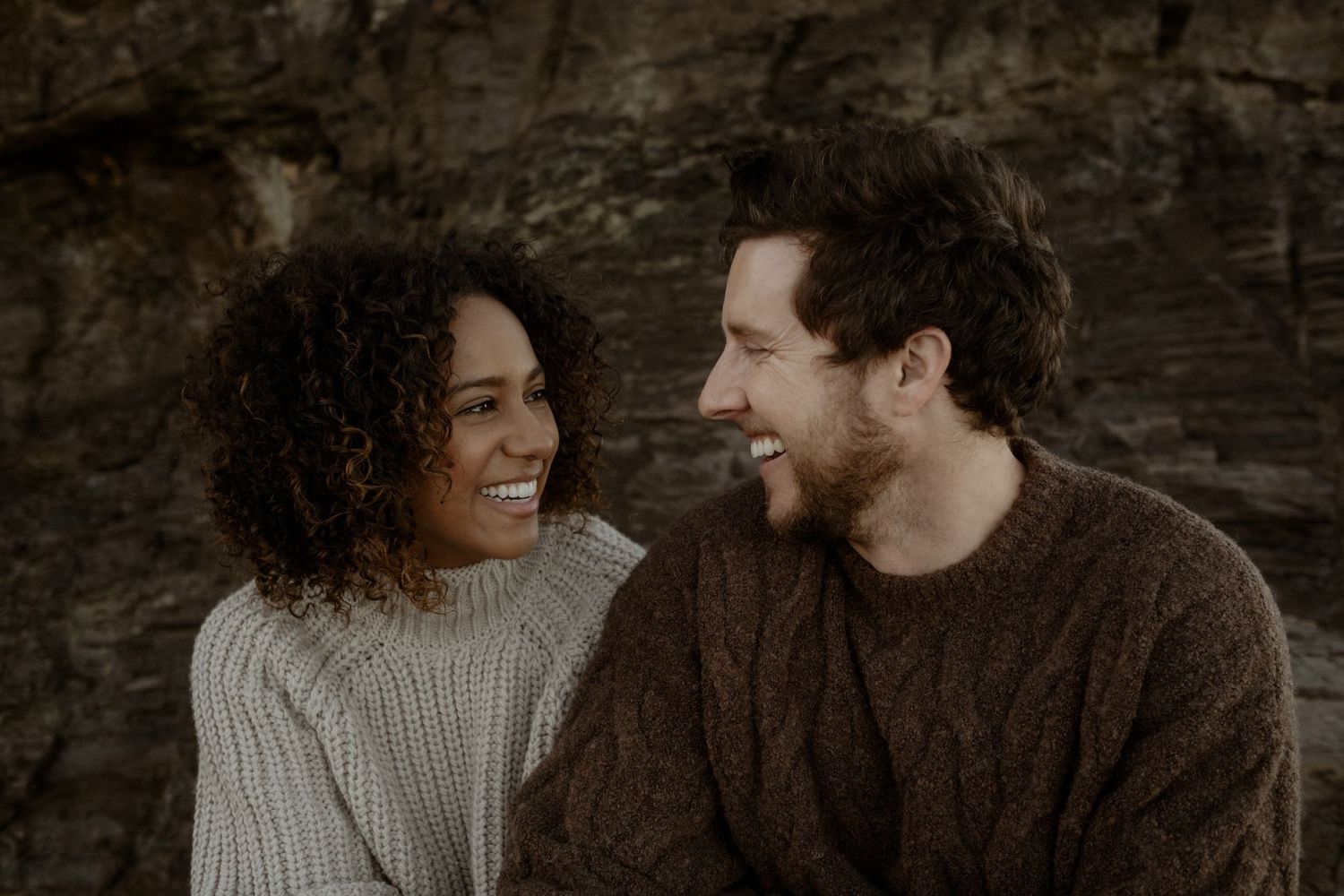 Couple sitting in front of a big rock smiling at each other.