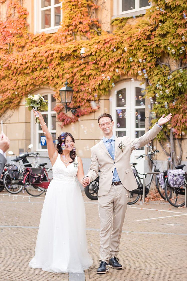 A wedding couple shares a joyful moment in front of an autumn-covered brick building after town hall wedding in Gorinchem with soap bubbles.