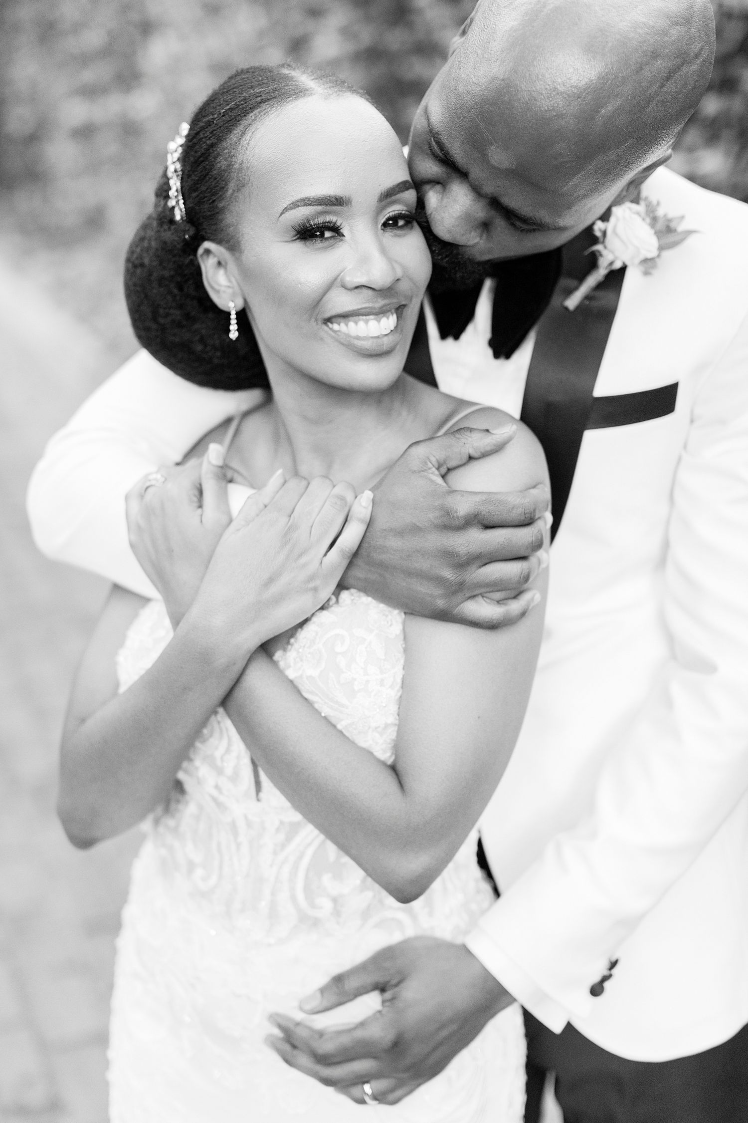 Black and white portrait of smiling bride in lace gown as groom tenderly kisses her cheek.