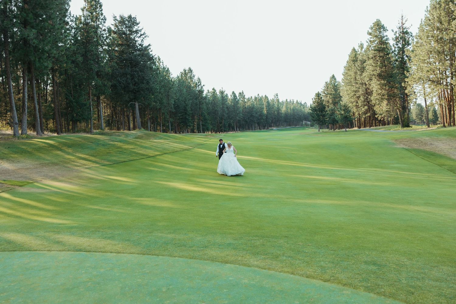 Wedding couple walking on manicured green golf course surrounded by tall pine trees.