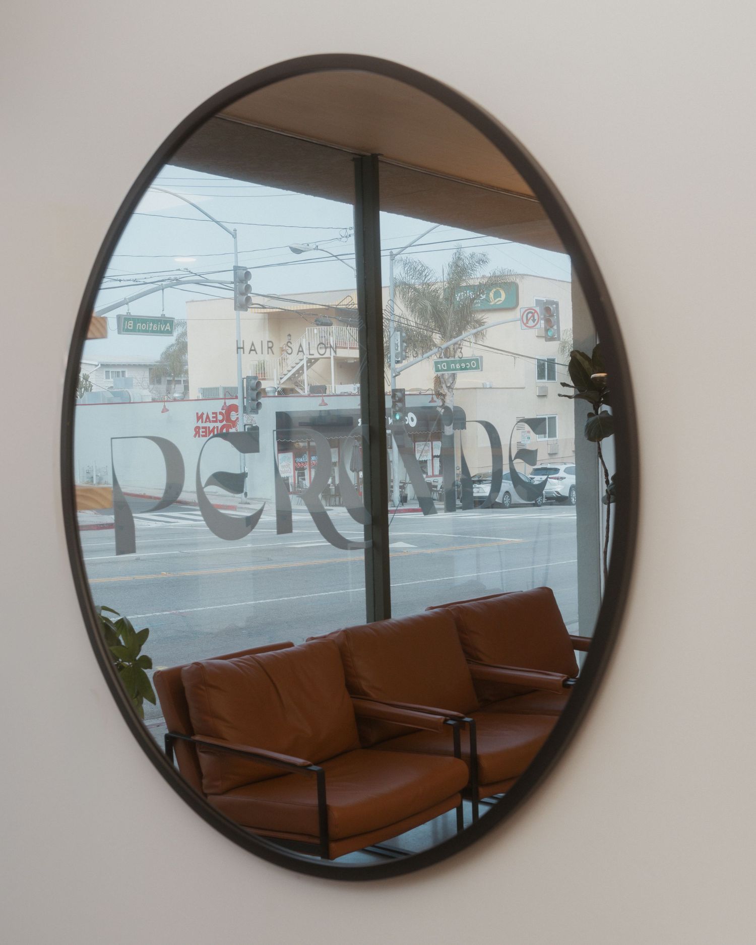 A brown leather couch is reflected in an oval wall mirror showing a storefront window with street view beyond.