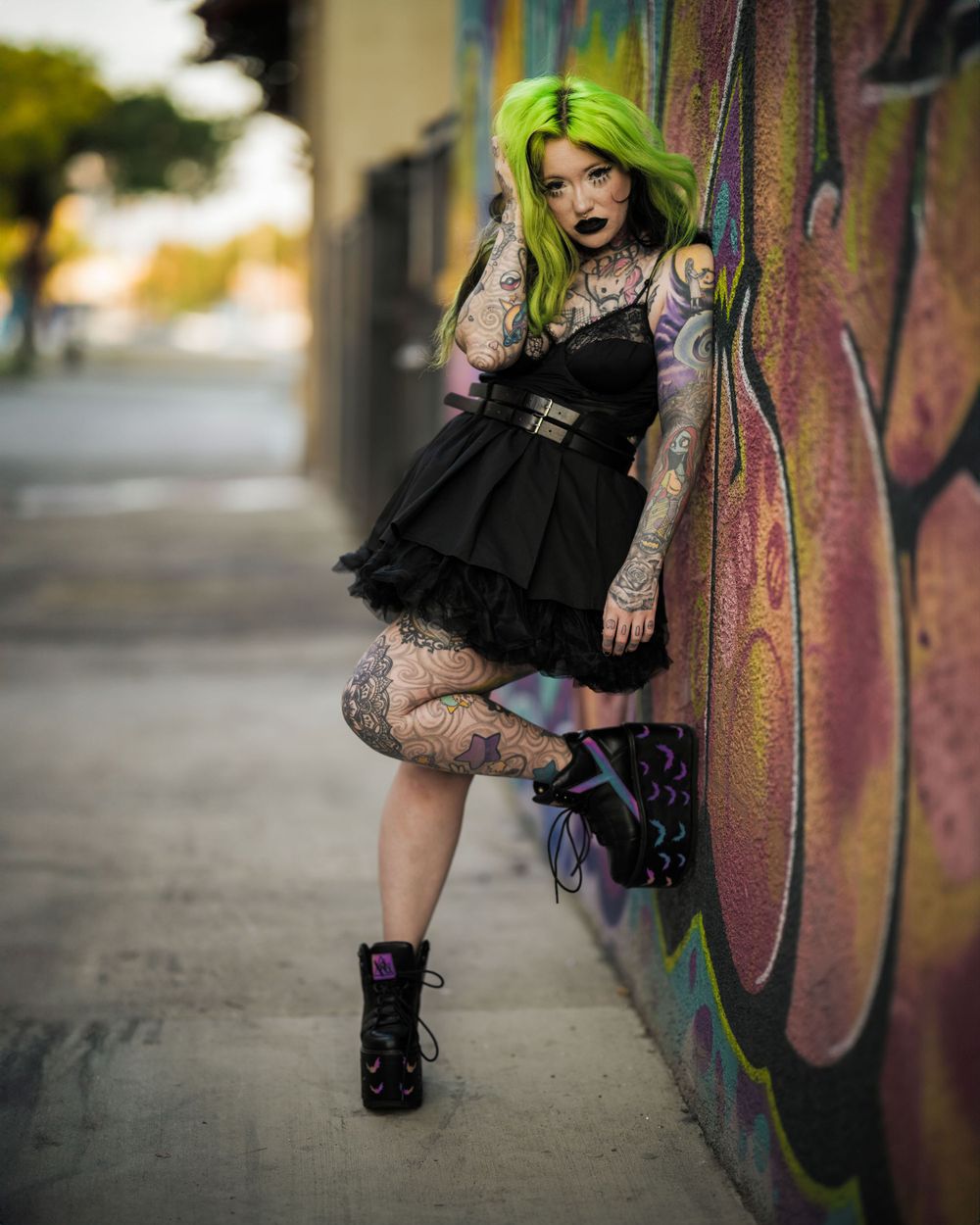 Heavily tattooed alt female model in a black dress and large black boots, posing against graffiti walls in Santa Ana's arts district
