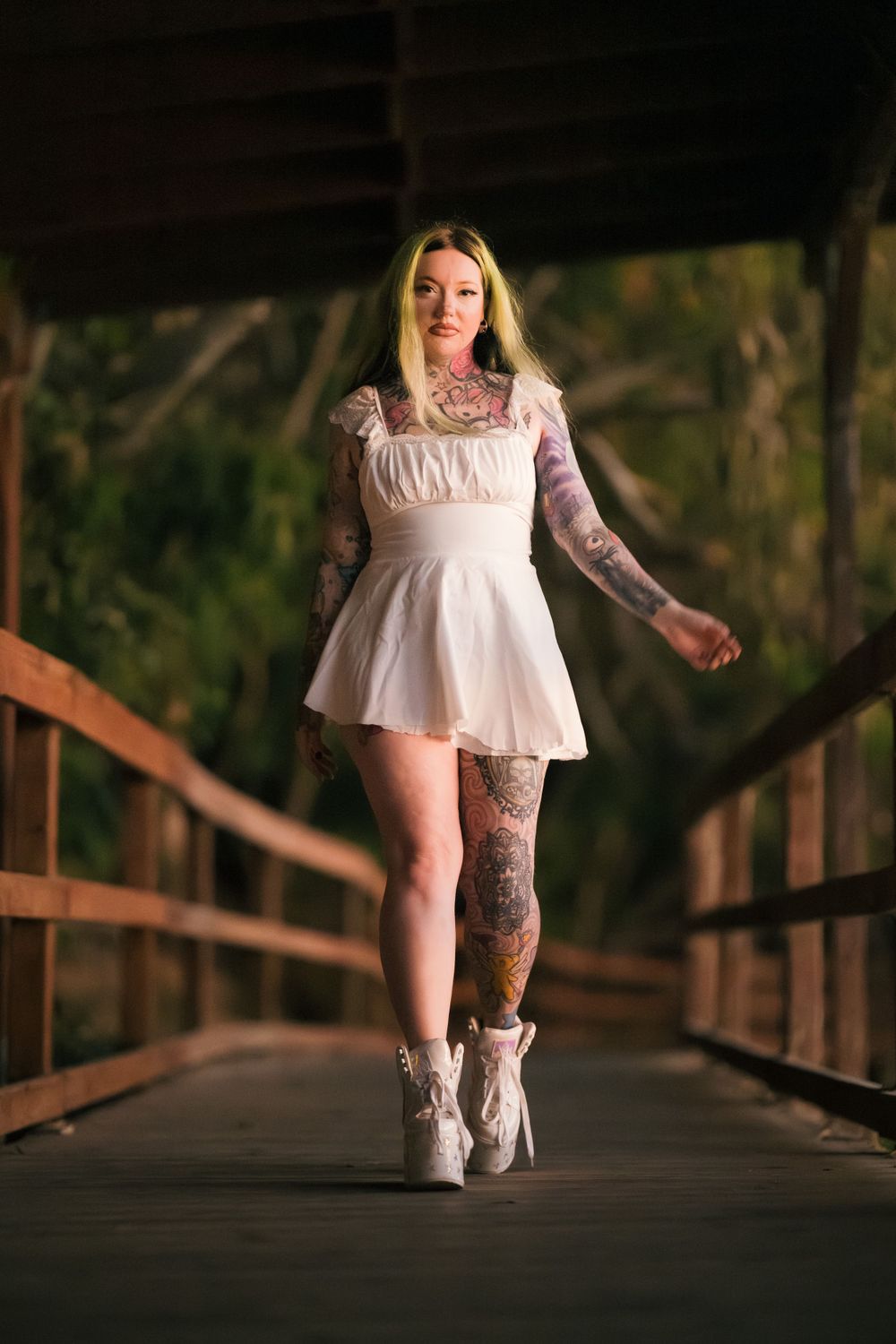 Heavily tattooed female model in a white dress on a wooden bridge by a lake, photographed just after sunset.