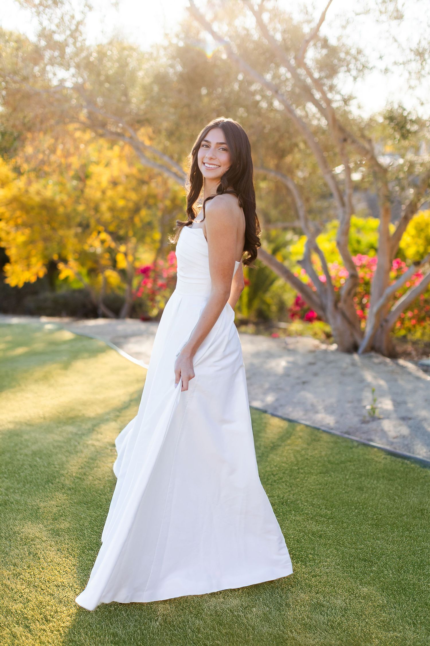 A high school senior wearing a flowing white dress glimmers in golden sunset light along a coastal path with palm trees and ocean views.