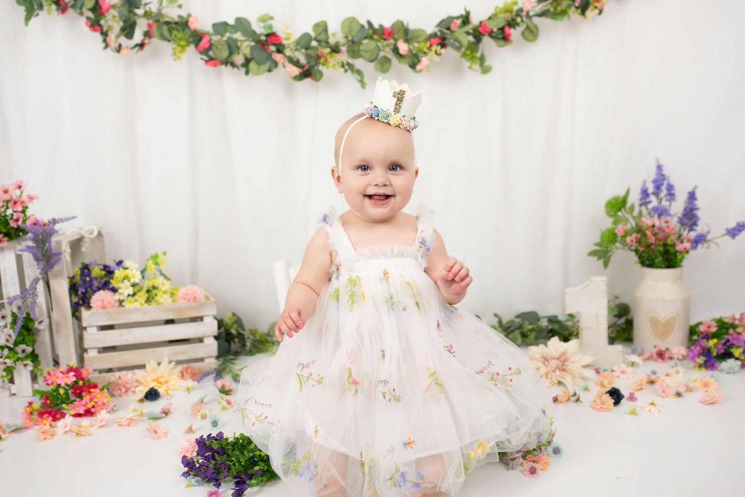 Smiling baby girl in white floral dress with gold crown headband, surrounded by colorful flowers and garlands.