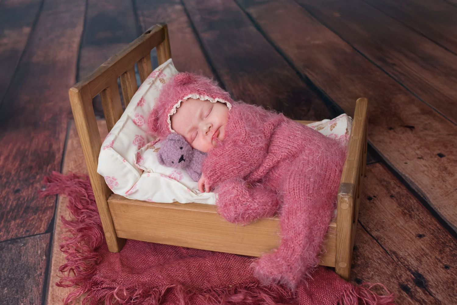 Newborn baby in pink fuzzy outfit sleeping in tiny wooden bed with stuffed animal on rustic wood floor.
