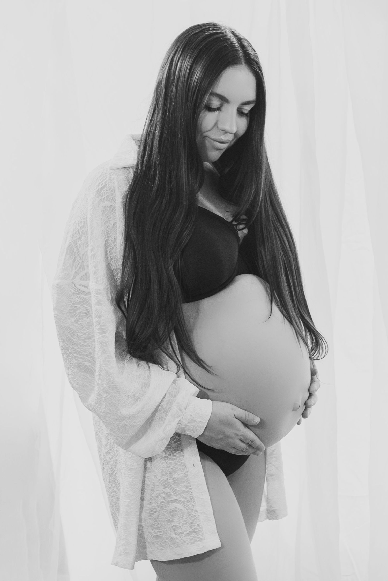 Black and white maternity photo of a pregnant woman with long dark hair cradling her baby bump in a lace robe.