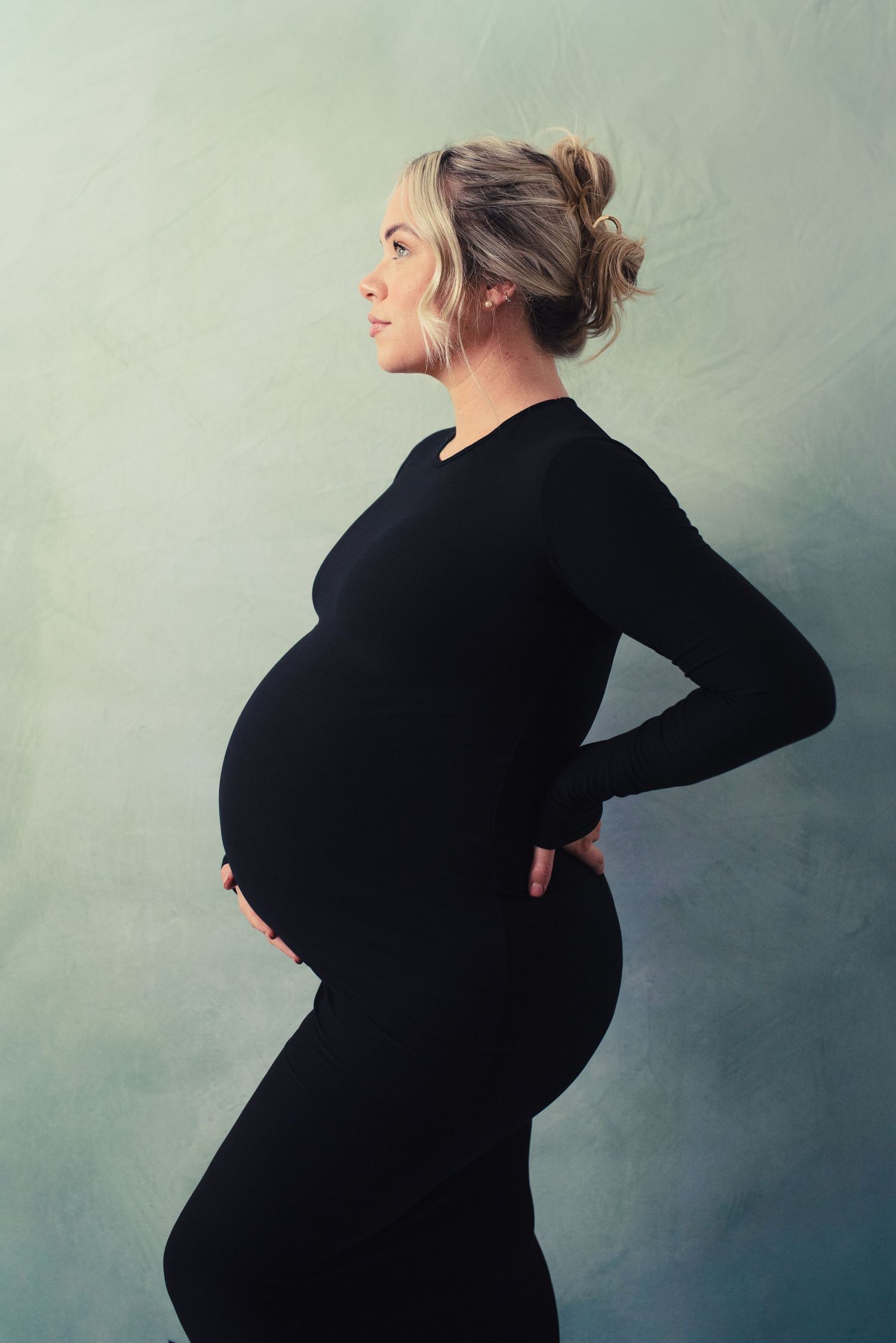 Pregnant woman in black outfit holding baby bump, side profile maternity photo on blue-gray background.