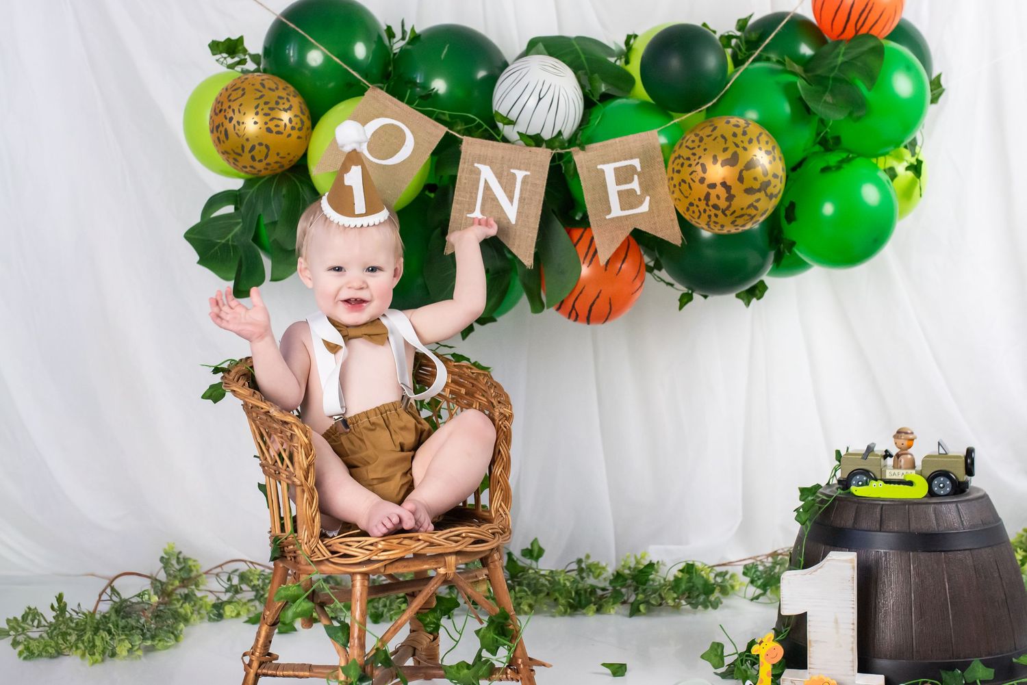 Baby's first birthday cake smash photo with green and gold balloon arch, ONE banner, and jungle theme decorations.