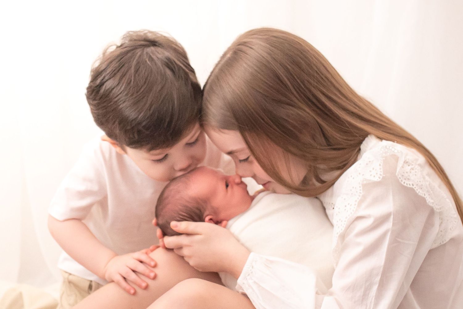 Two children in white clothing lovingly kiss a sleeping newborn baby in a bright, soft-lit portrait.