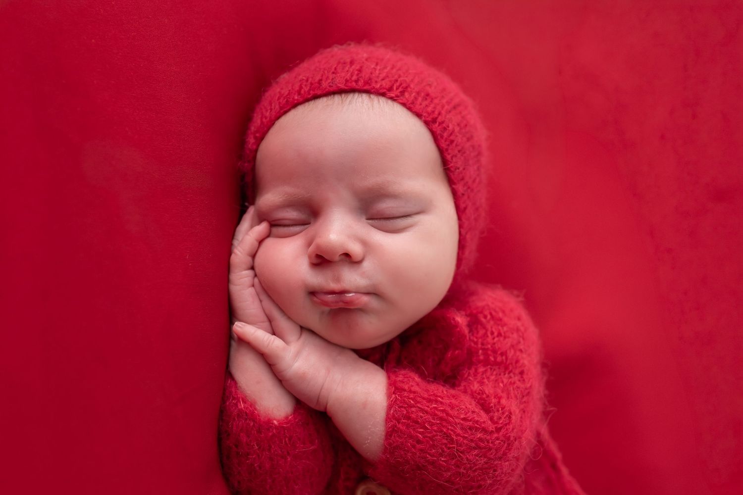 Newborn baby sleeping peacefully in red knit outfit and hat on red background.