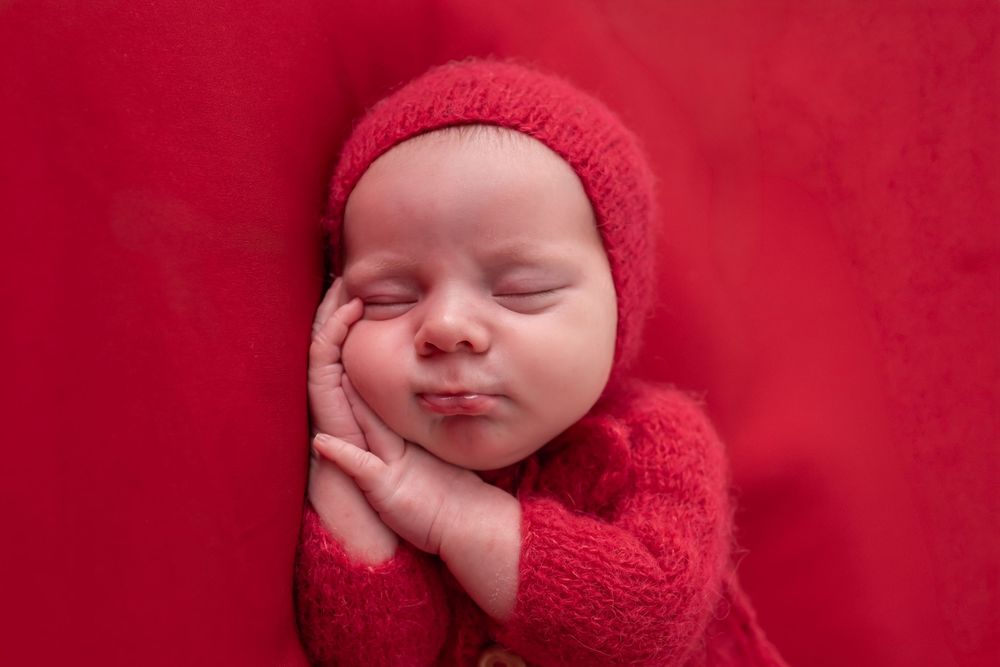 Newborn baby sleeping peacefully in red knit outfit and hat on red background.