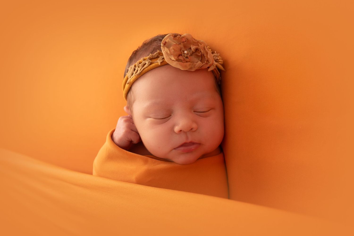Newborn baby swaddled in orange wrap with floral headband, sleeping peacefully on orange background.