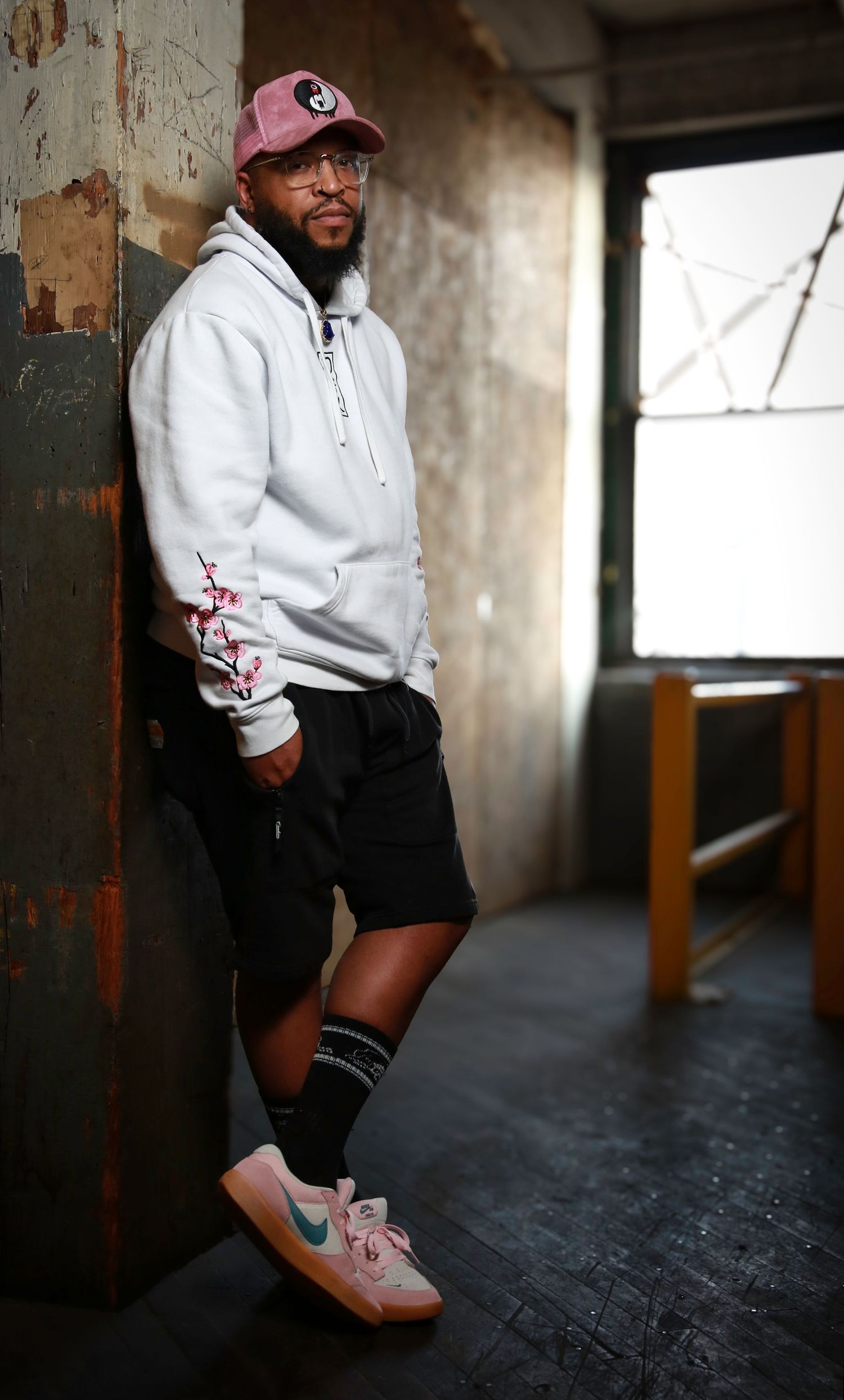 Person in white hoodie and black shorts poses against industrial backdrop wearing pink sneakers.