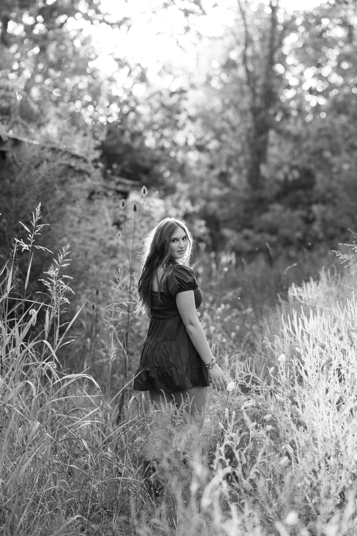 Black and white portrait of someone in a dress standing in tall grass at sunset.