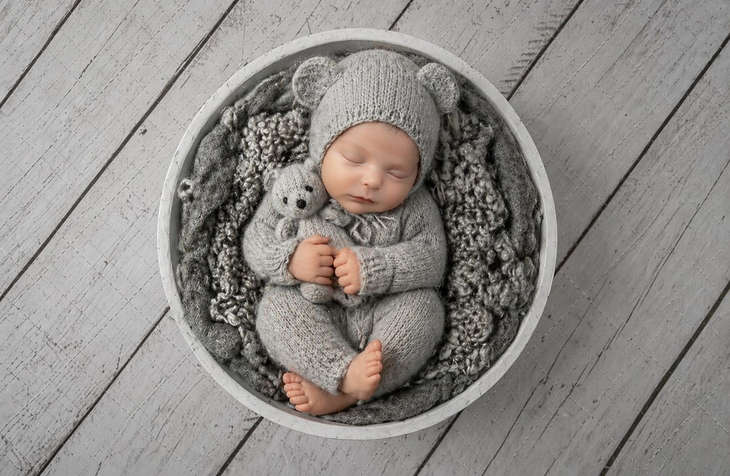 Newborn baby sleeping in white bowl wearing gray knit bear outfit, holding matching teddy bear on wood floor.
