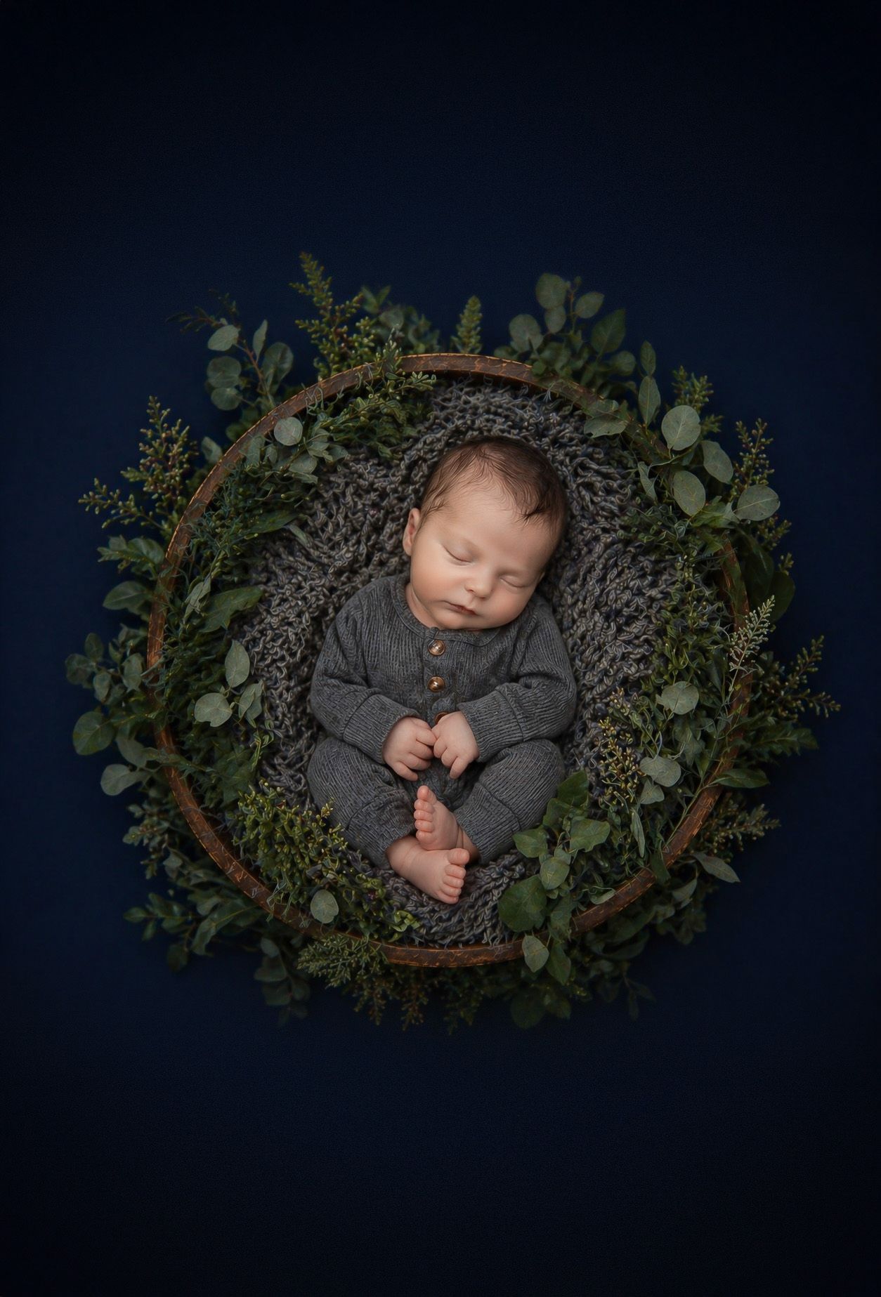 Newborn baby sleeping peacefully in a rustic wreath basket with lush greenery on a dark navy background.