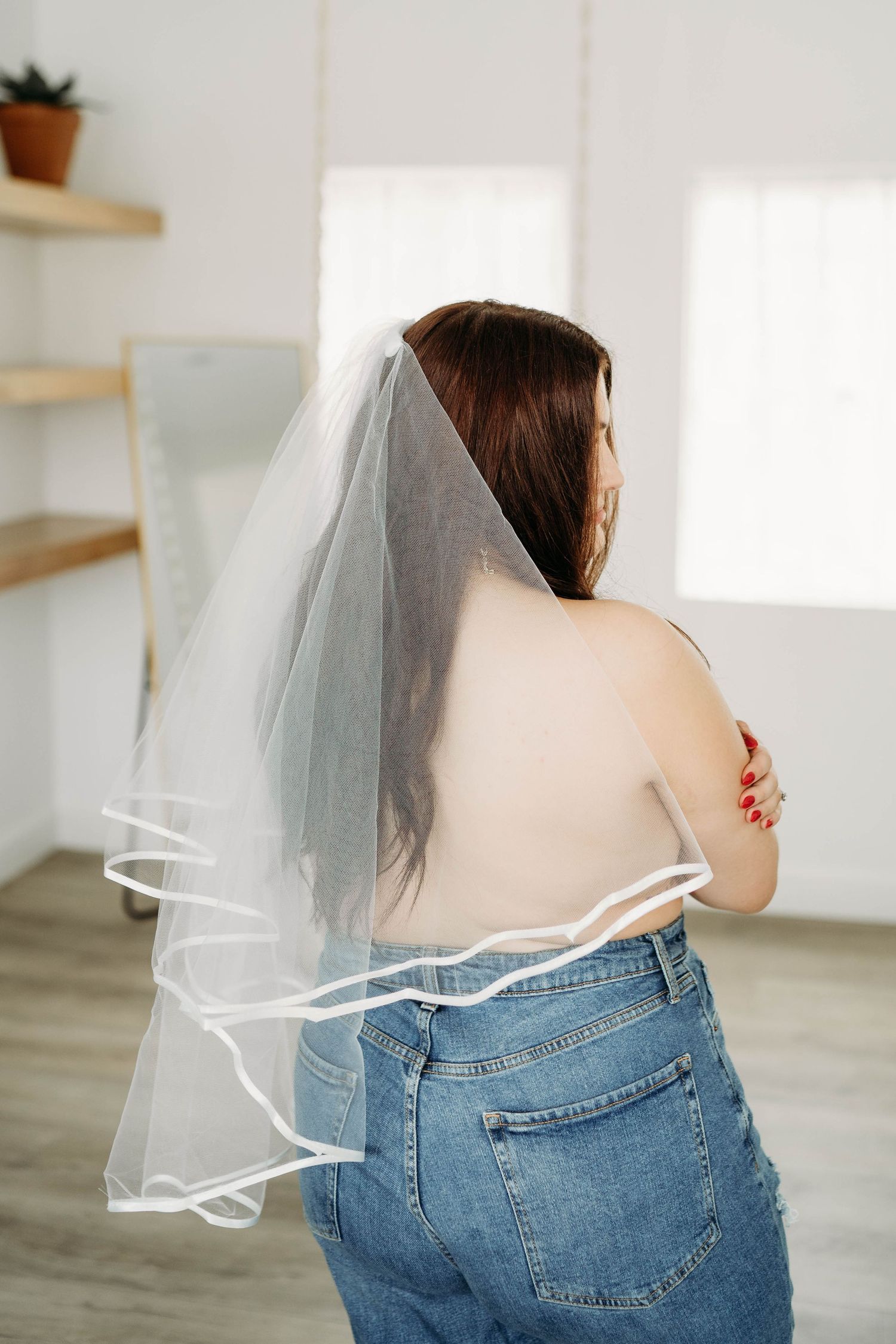 Back view of someone wearing a bridal veil and jeans in a bright minimal room.