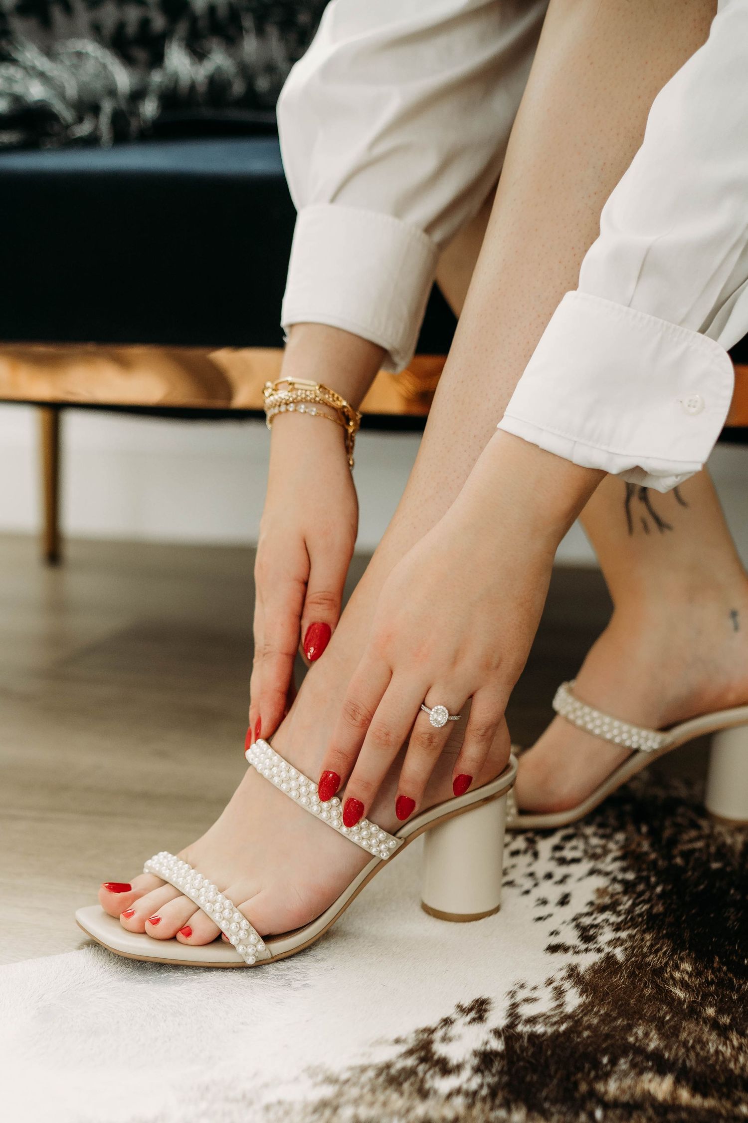 Person's hand with red nail polish adjusts pearl-embellished white heeled sandals while wearing white pants.