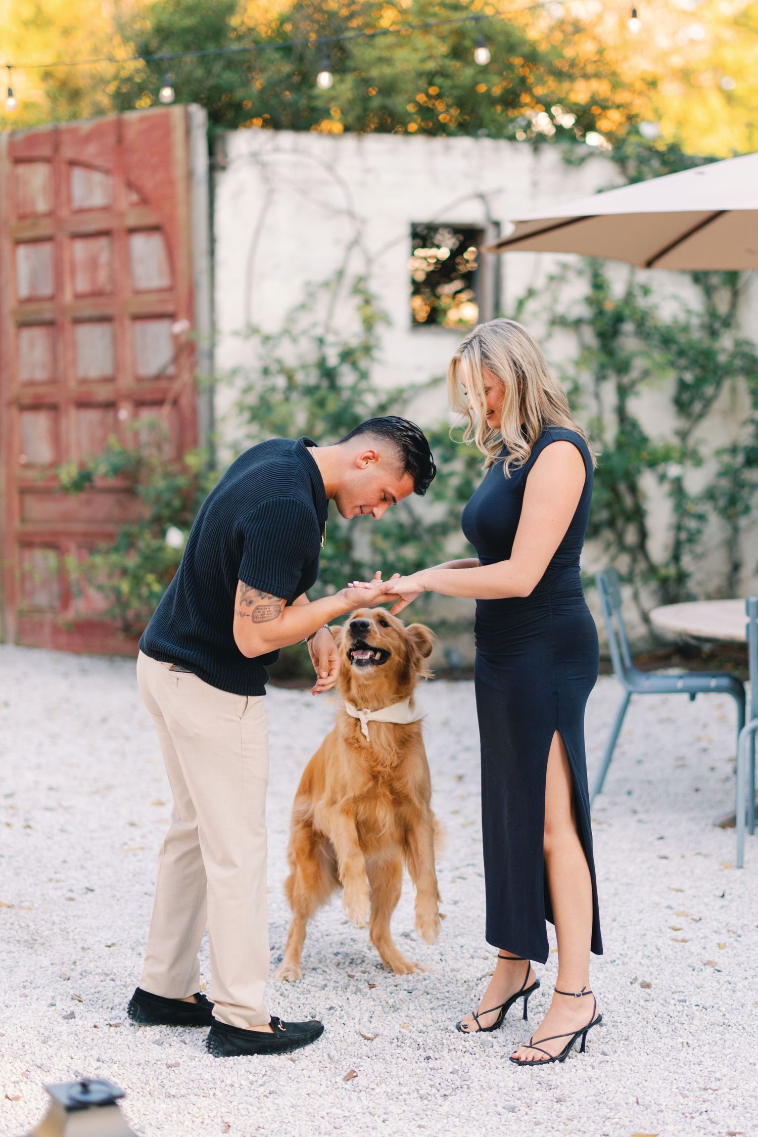 Couple in navy outfits petting their happy golden retriever dog in a charming outdoor patio setting at golden hour.