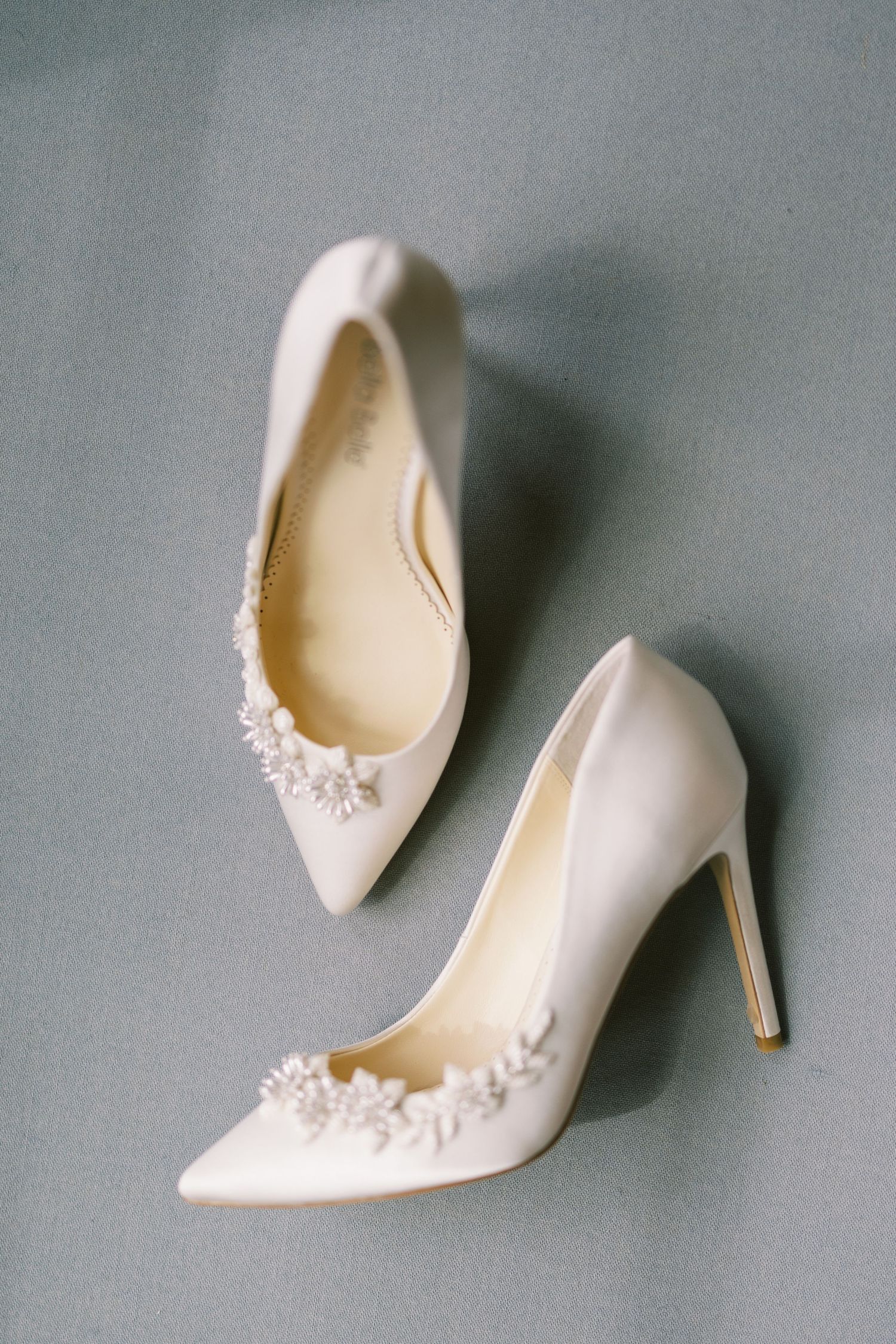 Elegant white bridal heels with delicate floral embellishments on pointed toe stilettos displayed on gray background.