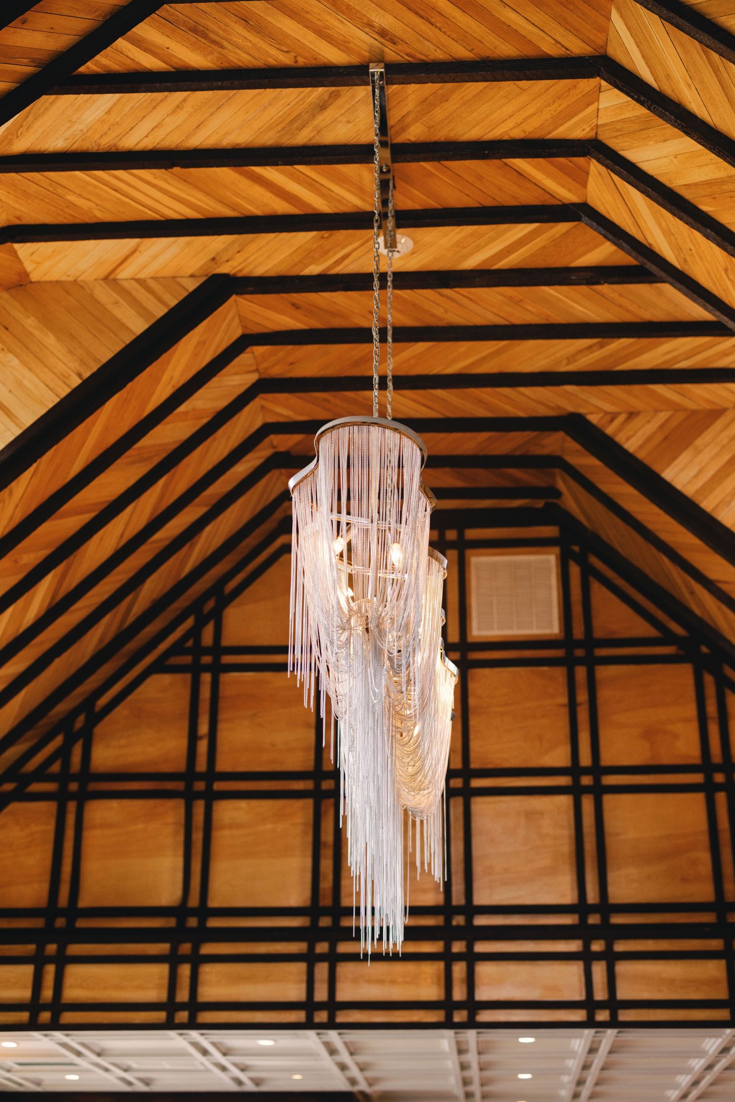 Elegant cascading crystal chandelier hanging from a vaulted wooden ceiling with geometric black beam detailing.