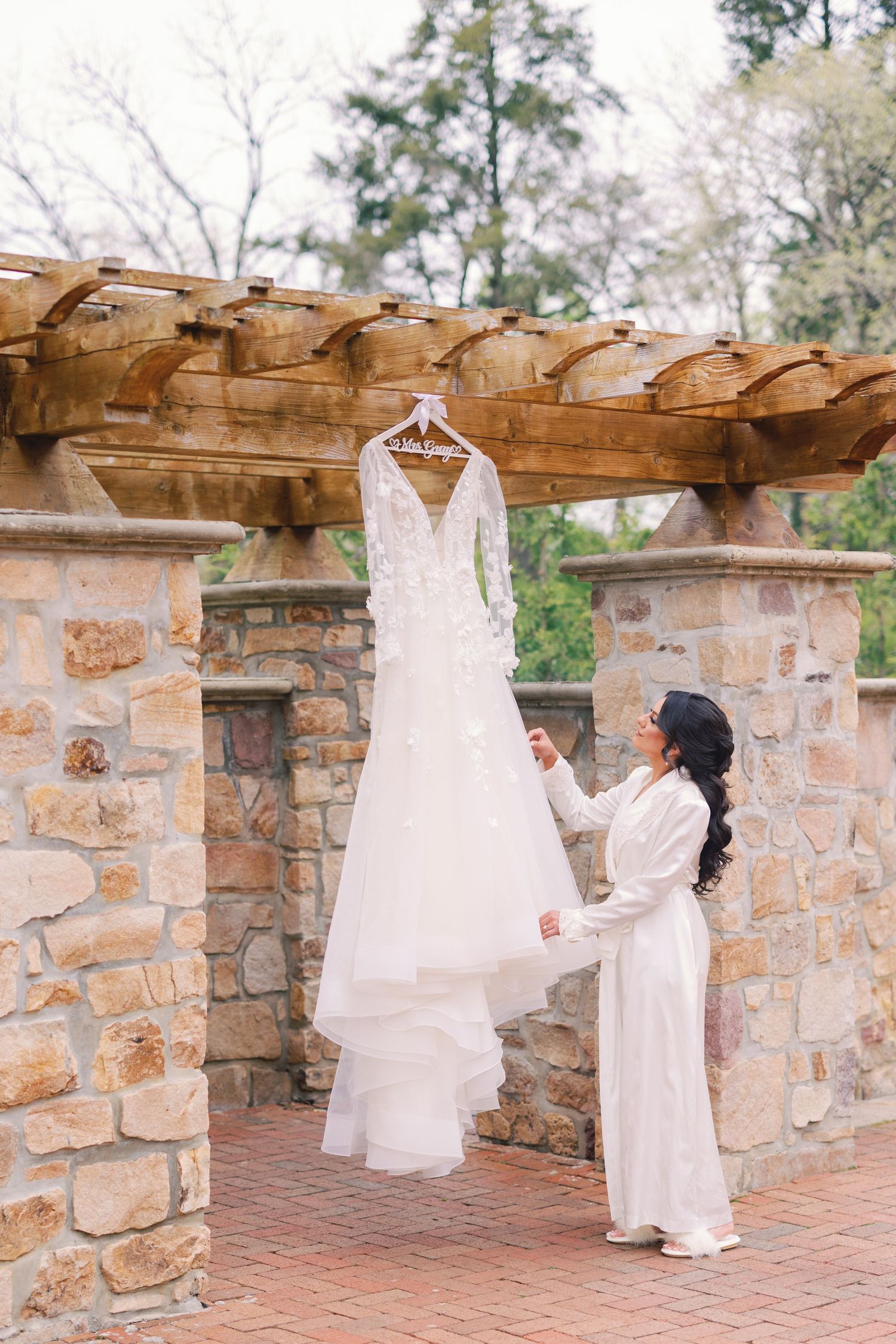 Bride in white robe admiring her lace wedding gown hanging under a rustic stone pergola outdoors.