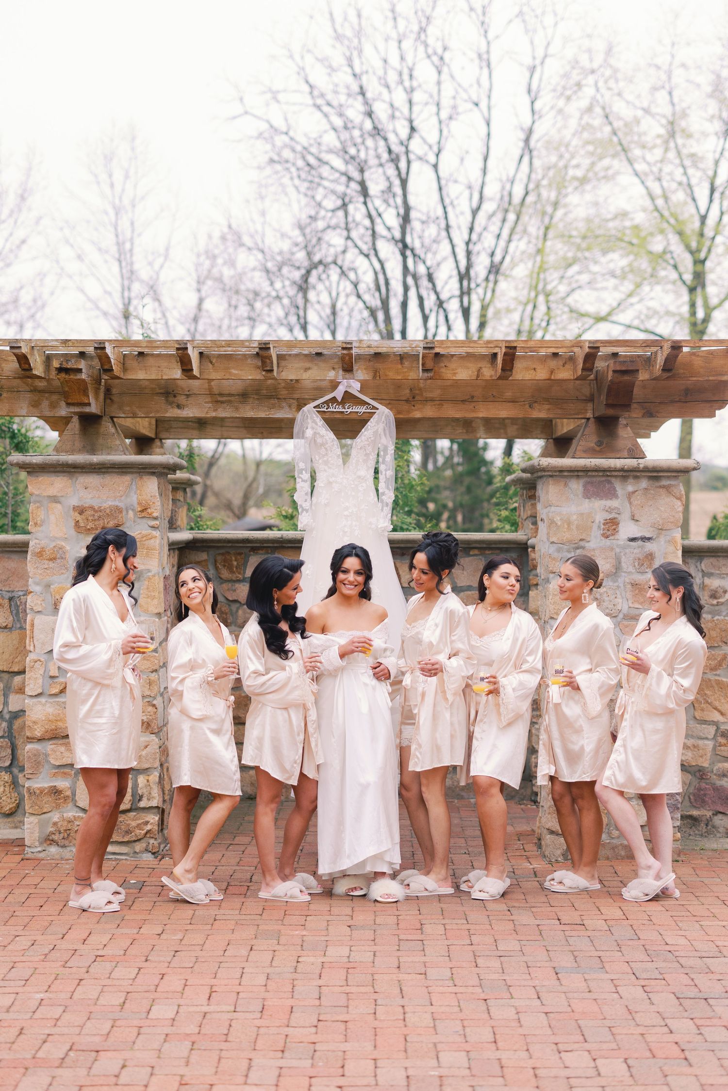 Bride in white robe poses with seven bridesmaids in champagne robes beneath hanging wedding dress on brick patio.