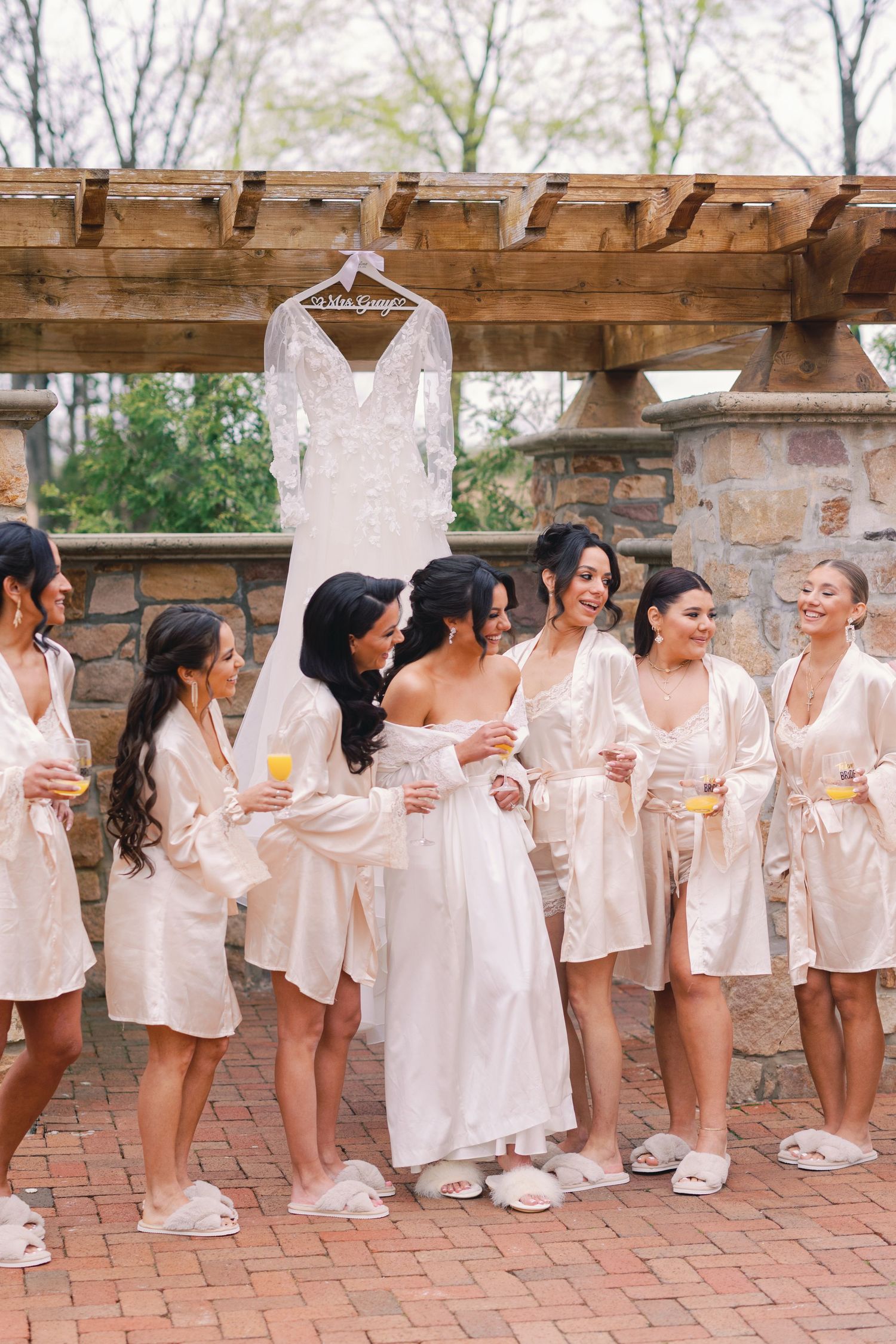 Bride and bridesmaids in champagne satin robes holding mimosas beneath hanging lace wedding gown on stone patio.