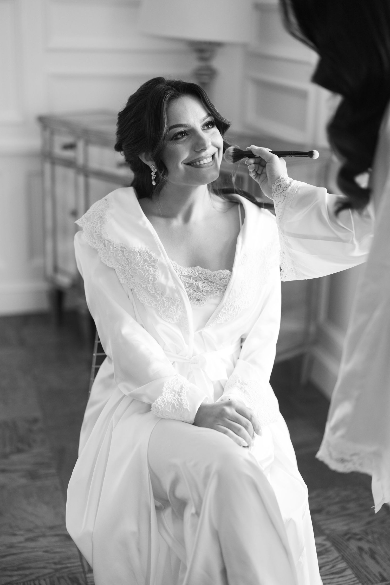 Black and white photo of smiling bride getting makeup applied while seated in white lace robe on wedding day.