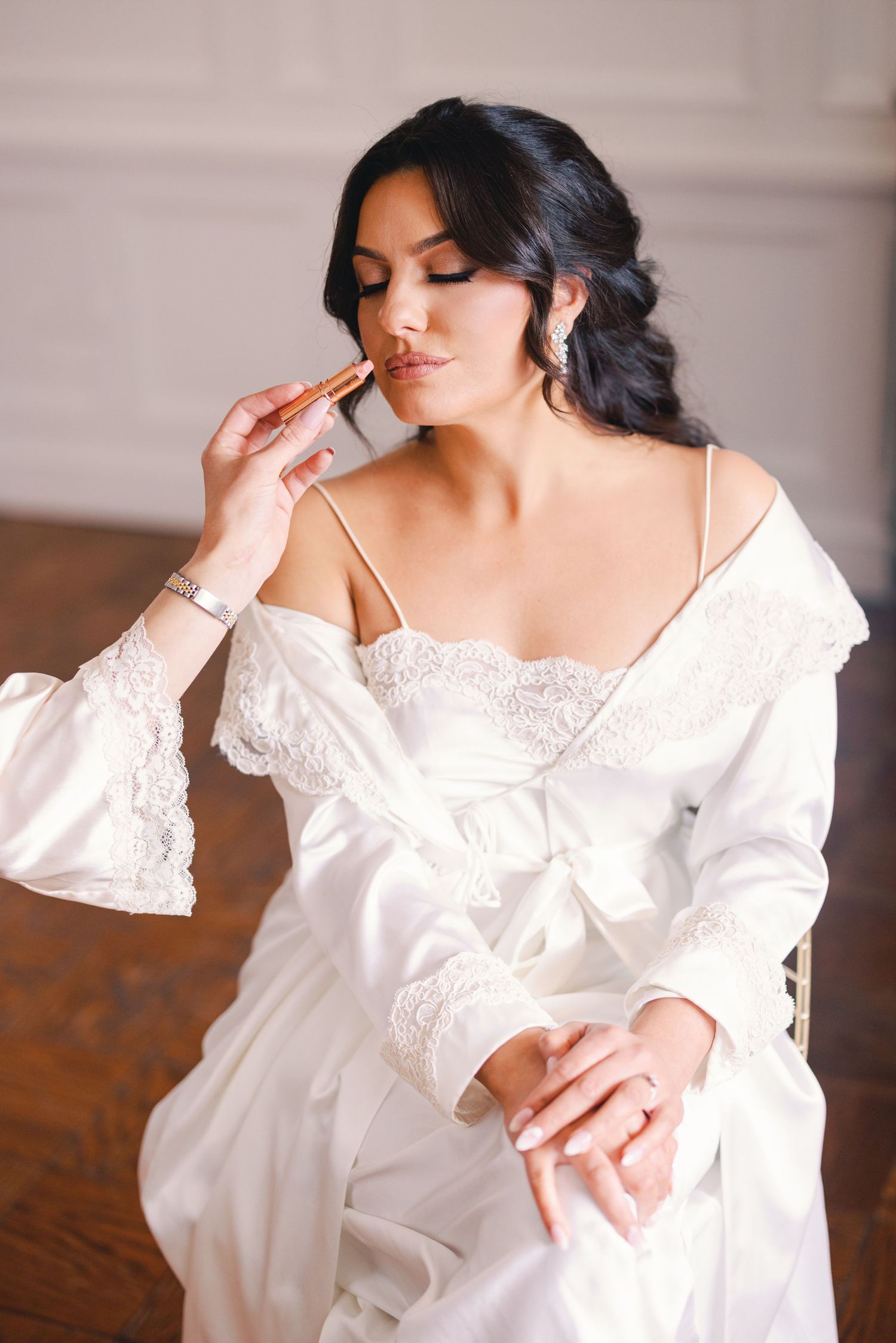 Bride in white satin lace robe applying makeup while seated, bridal morning preparation.