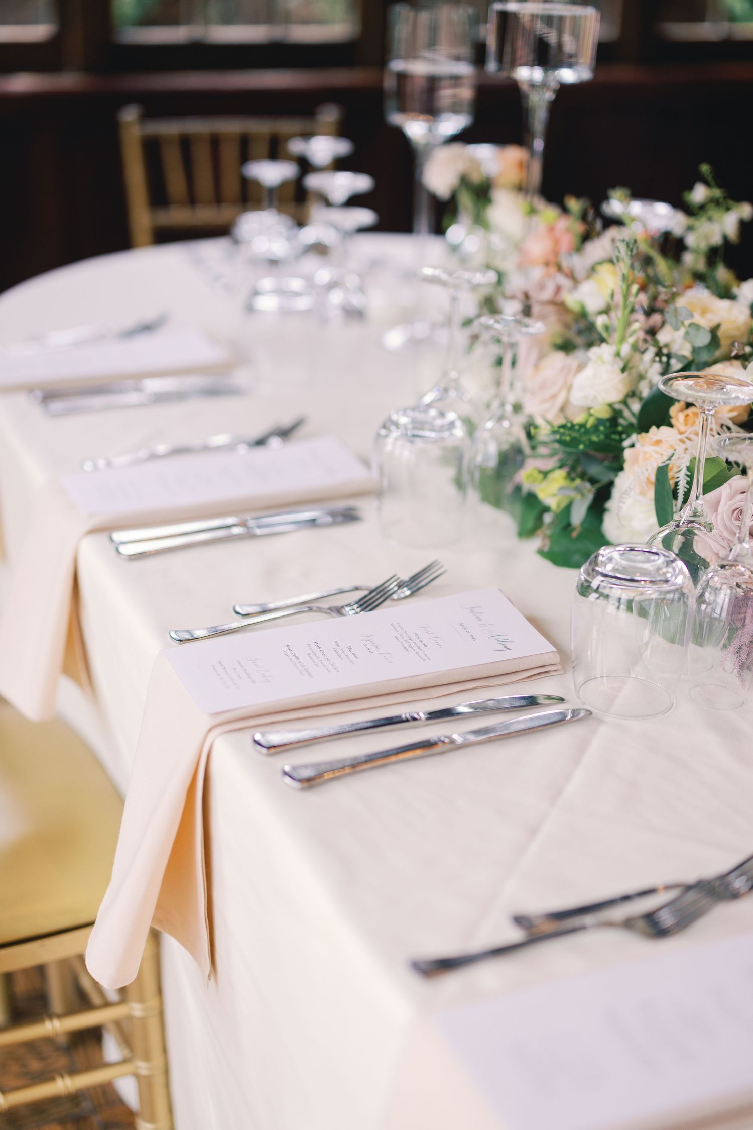 Elegant wedding reception table setting with blush napkins, silverware, menus, and floral centerpiece.