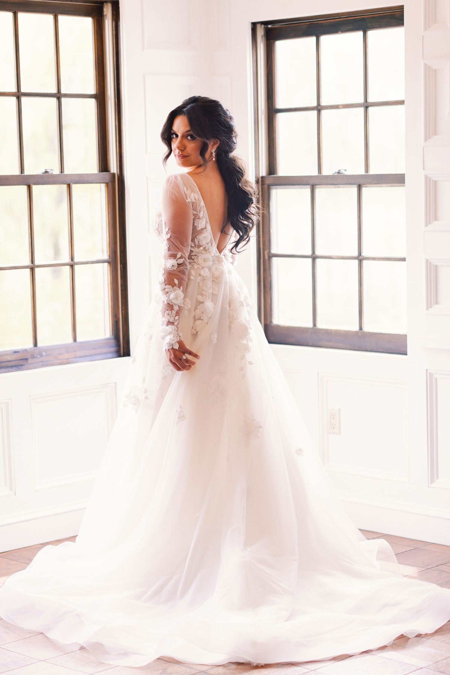 Elegant bride in floral lace long-sleeve wedding gown with open back standing by windows.