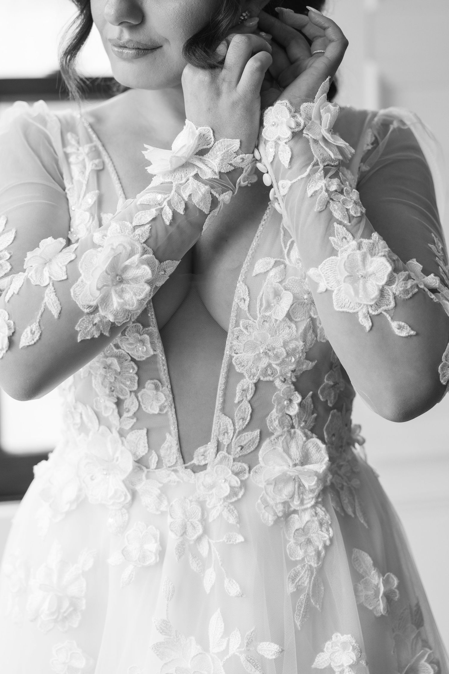 Close-up black and white detail of bride in floral lace wedding gown adjusting earring.