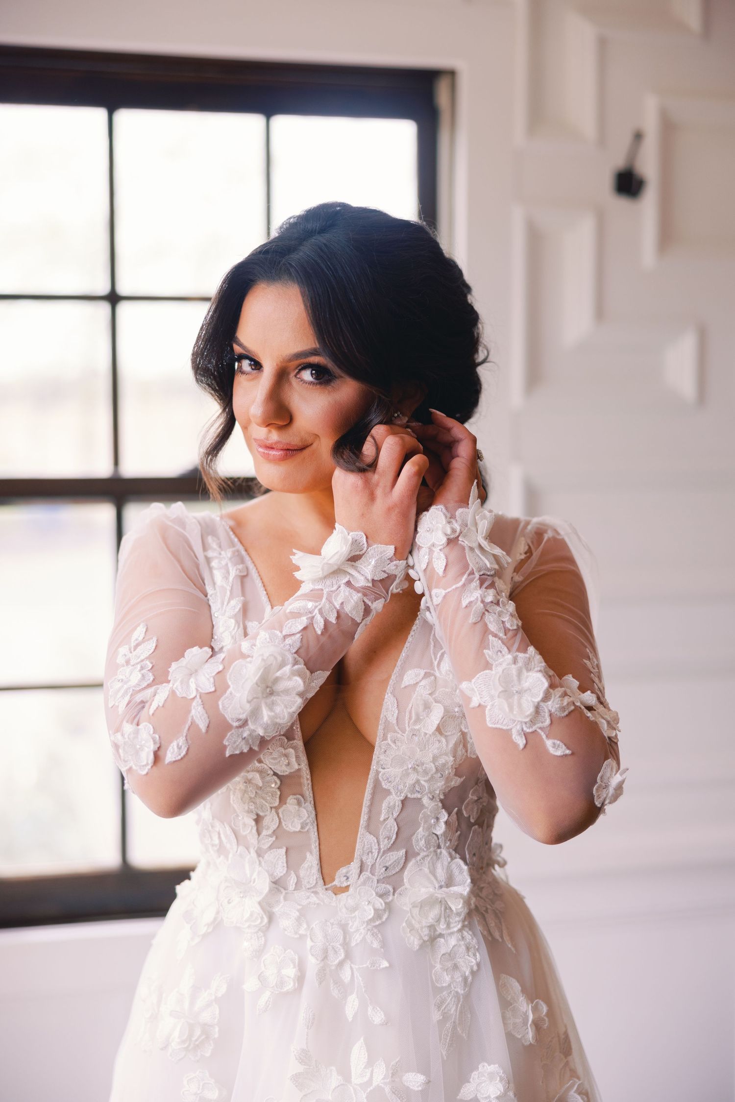 Dark-haired bride in floral lace deep V-neck wedding gown adjusting earring indoors.