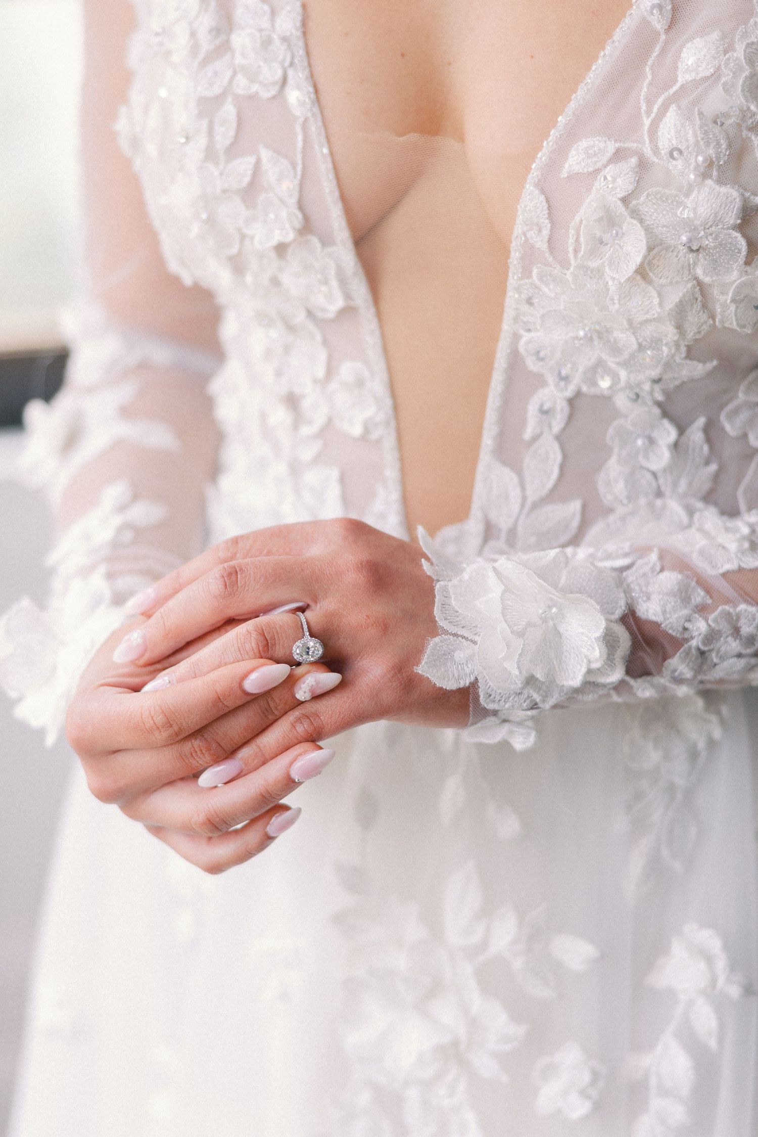 Close-up of bride\u2019s hands with oval diamond engagement ring wearing floral lace wedding gown.