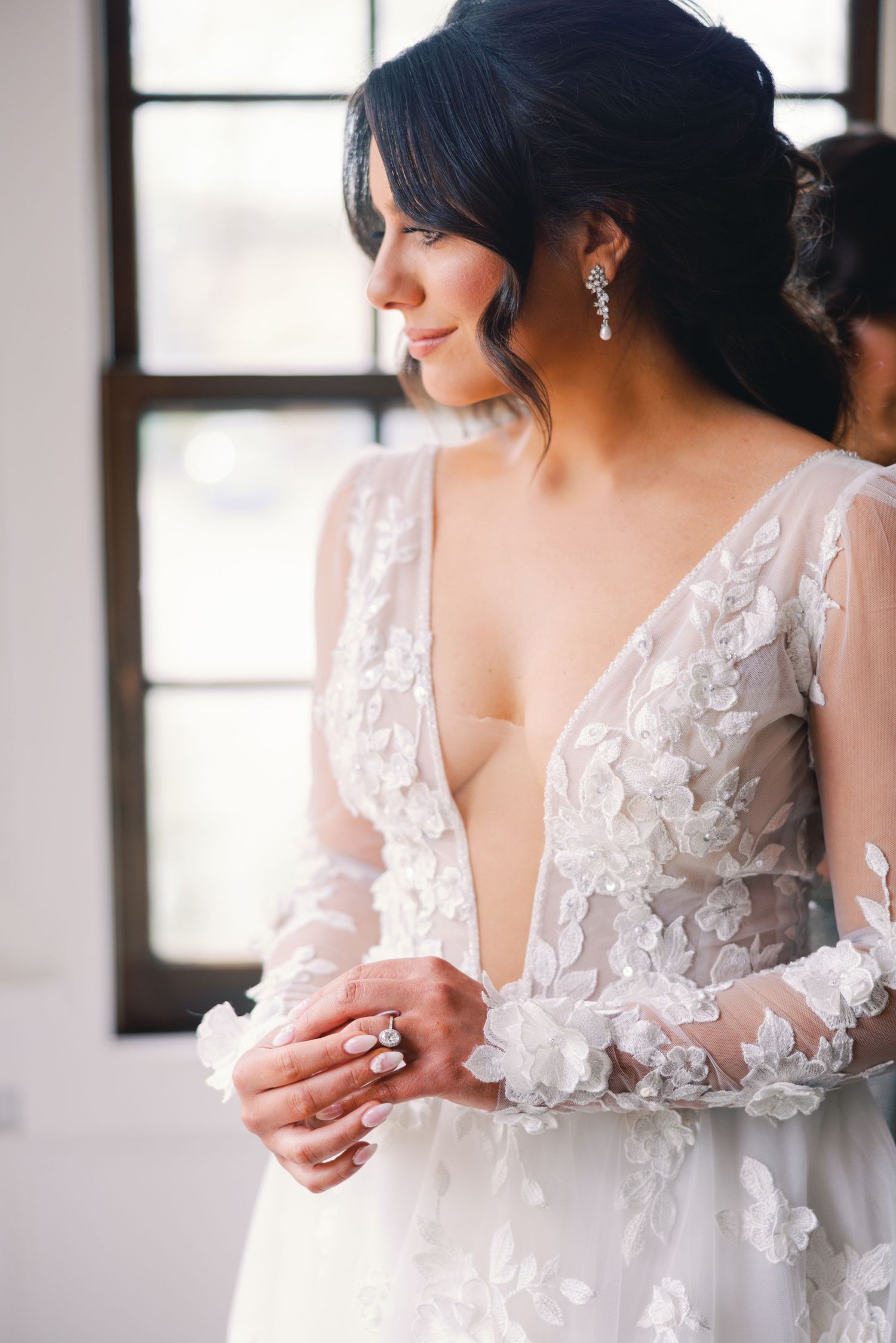 Bride in deep V-neck floral lace wedding gown with diamond ring and drop earrings.