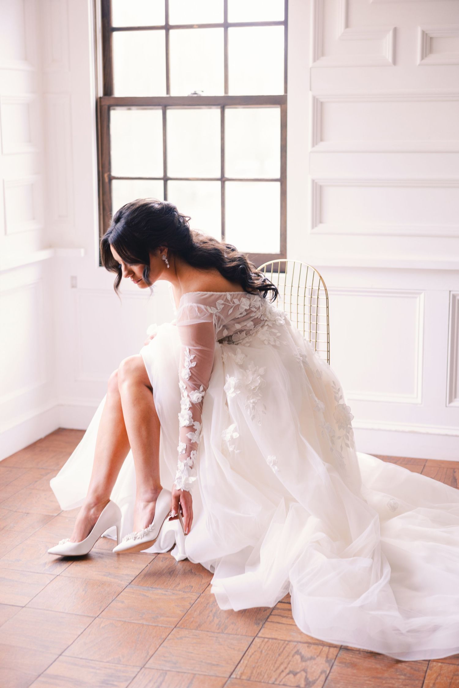 Bride in floral lace wedding gown putting on white heels while seated on floor.
