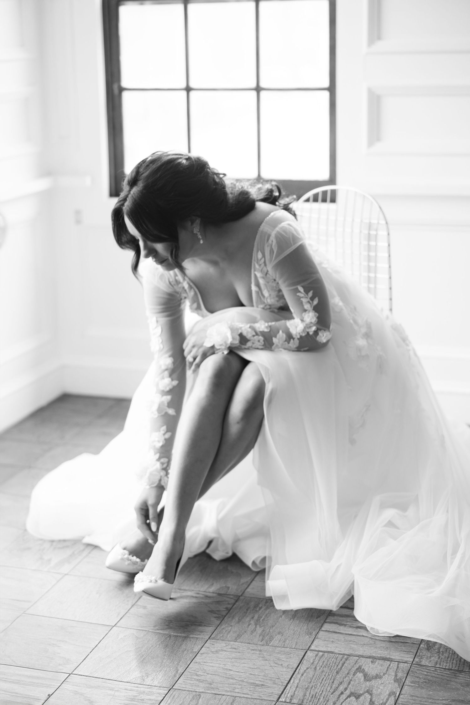 Black and white photo of bride in lace gown adjusting her heel while seated near a window.