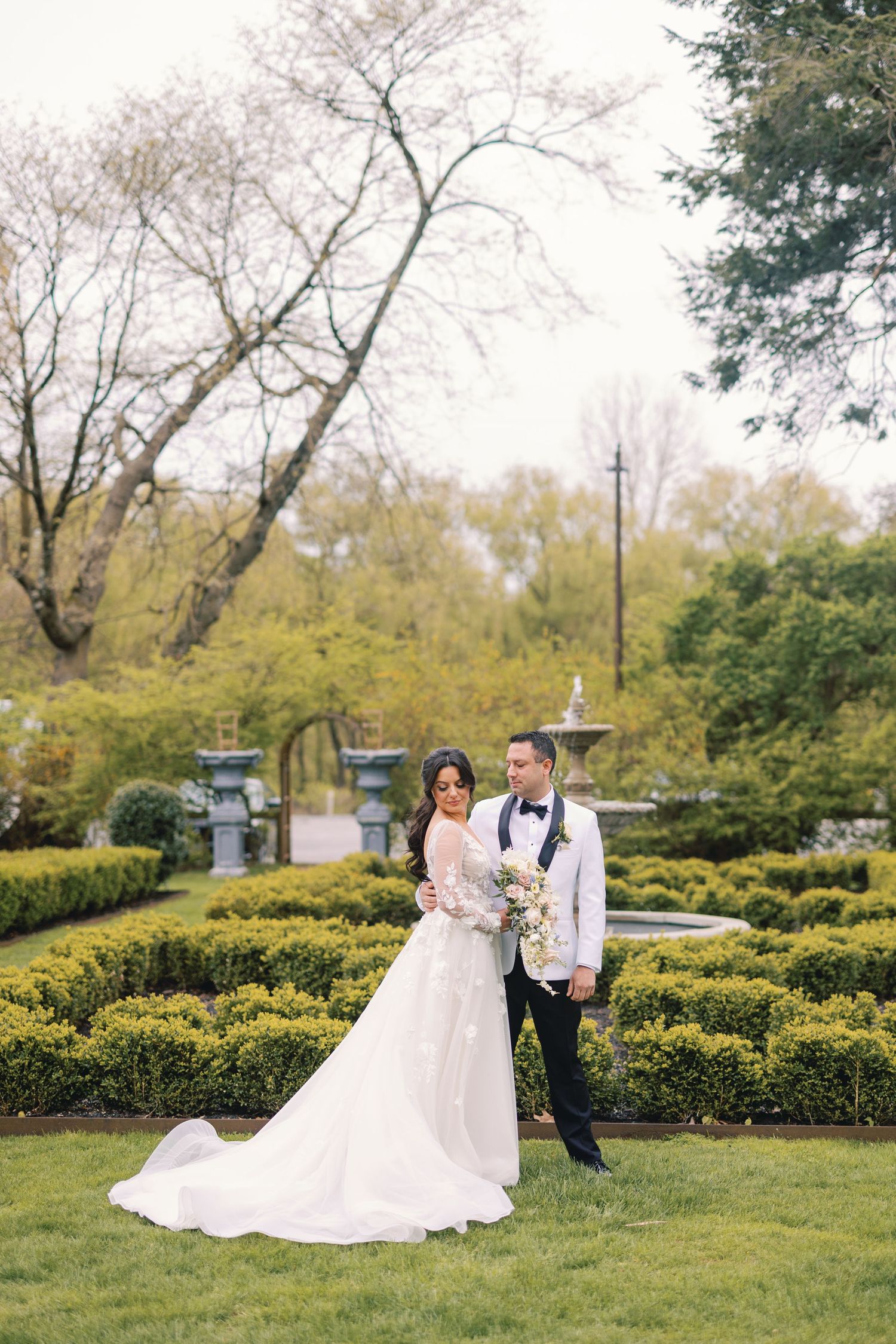 Elegant bride and groom posing in a formal garden with manicured hedges and fountain.