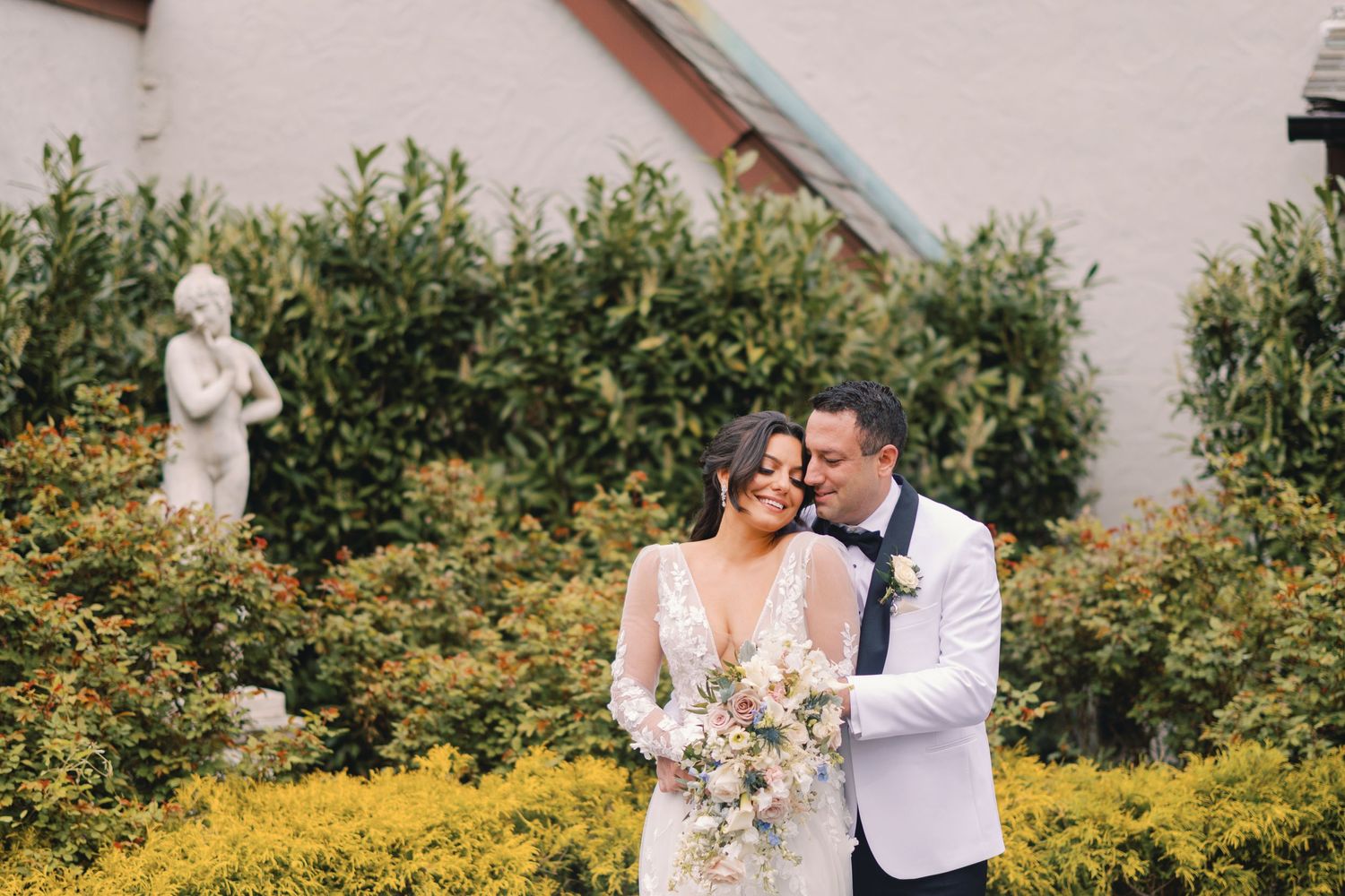 Bride and groom embracing near yellow bushes and a stone statue outdoors.