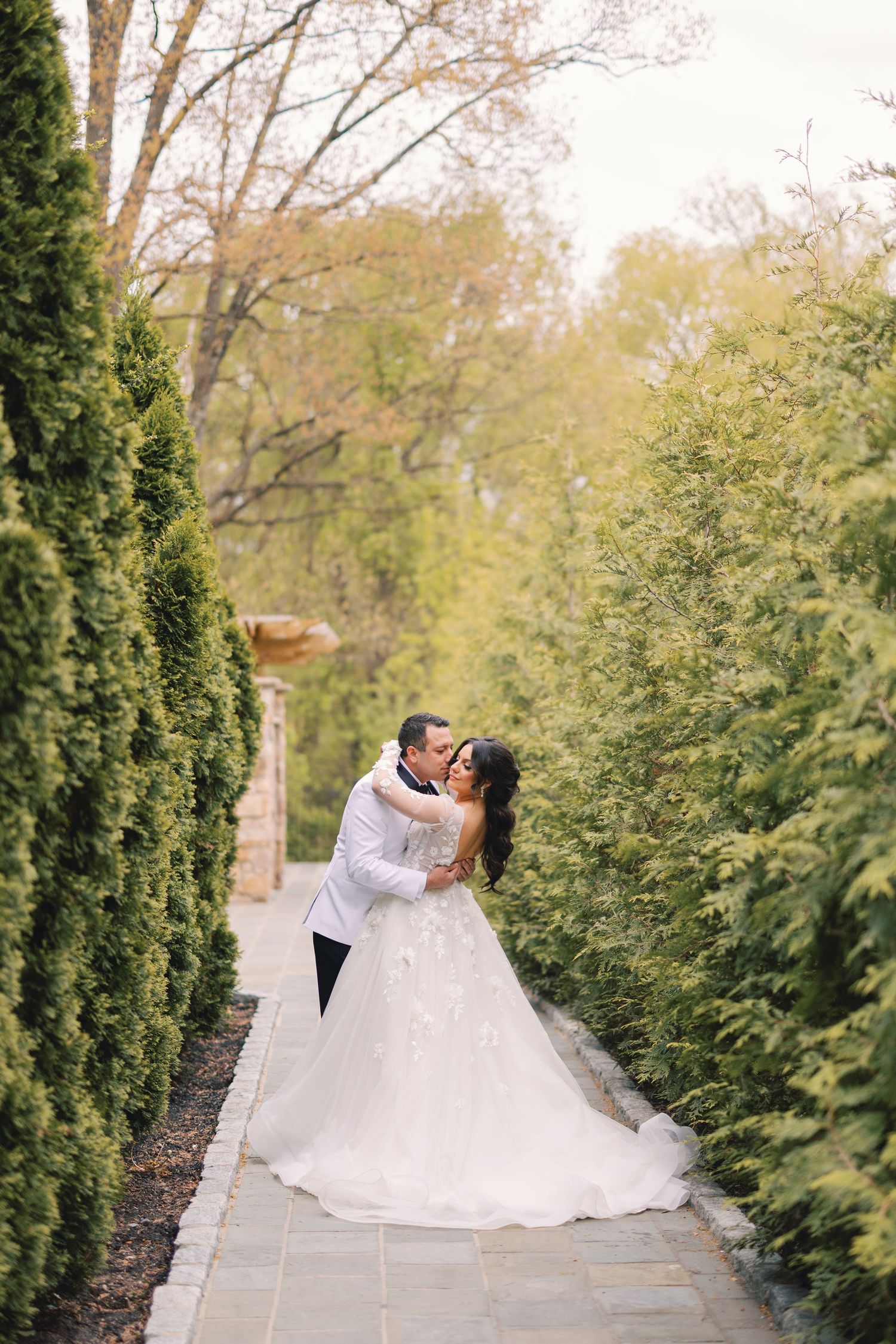 Bride and groom kissing on a stone pathway lined with tall green cypress trees.