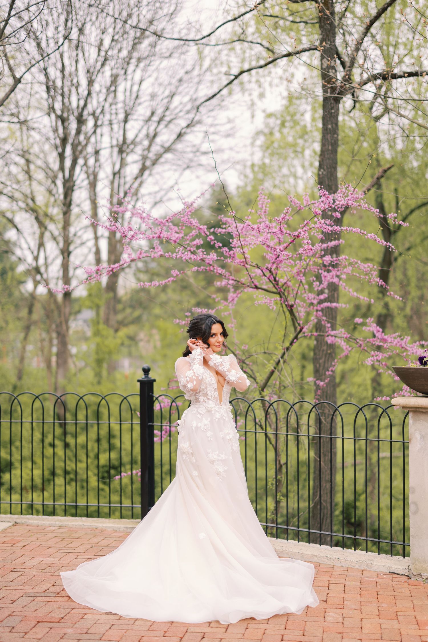 Bride in lace gown standing near a blooming pink cherry blossom tree outdoors.
