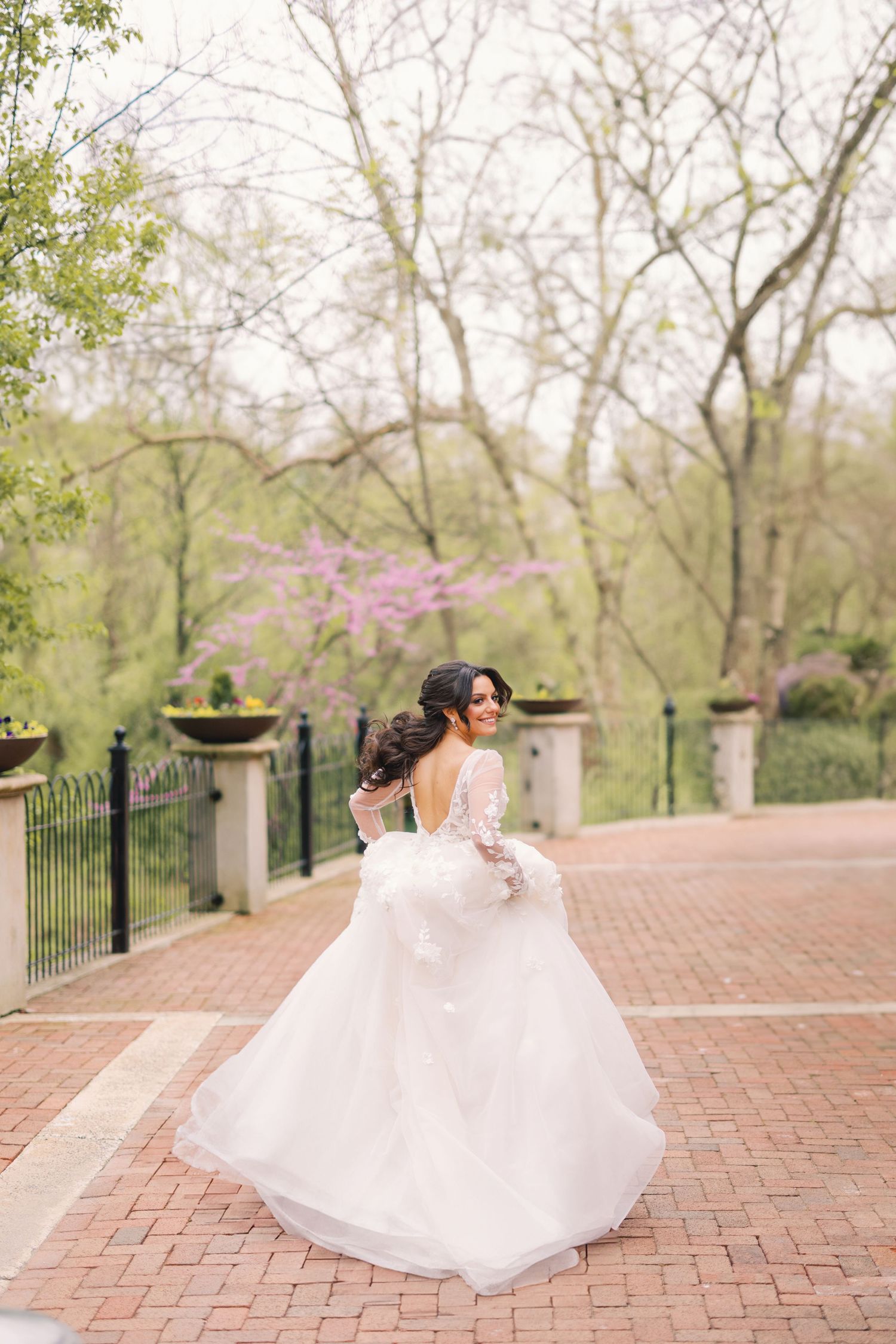 Smiling bride twirling in a flowing lace gown on a brick pathway surrounded by trees.